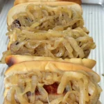 Four beer-braised bratwursts in toasted hoagie buns piled high with golden caramelized onions on a metal tray