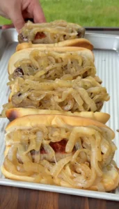 Four beer-braised bratwursts in toasted hoagie buns piled high with golden caramelized onions on a metal tray