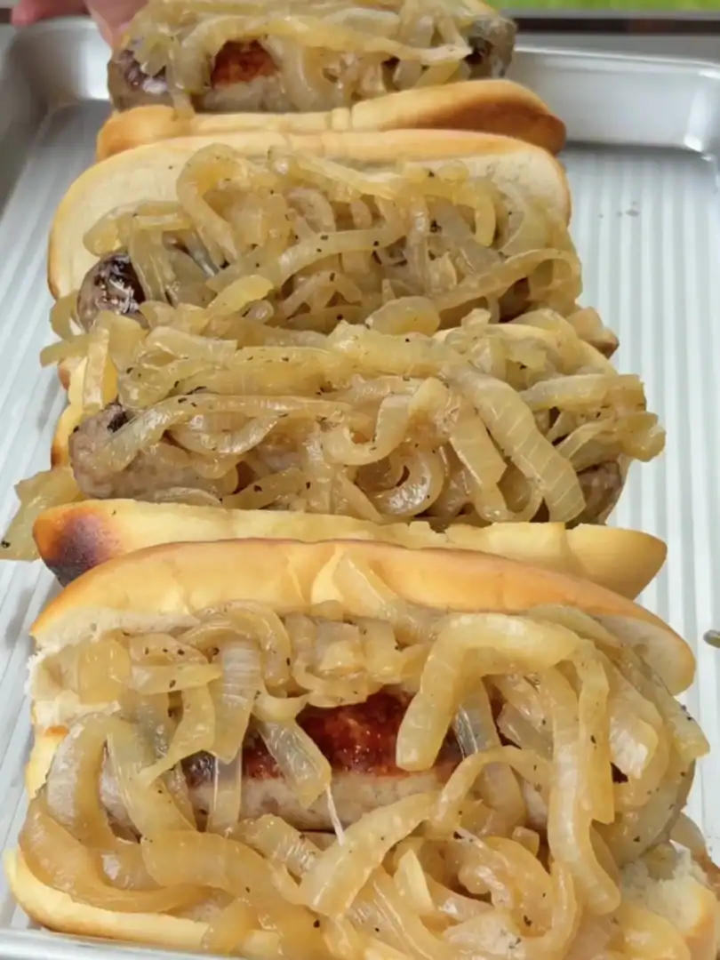 Four beer-braised bratwursts in toasted hoagie buns piled high with golden caramelized onions on a metal tray