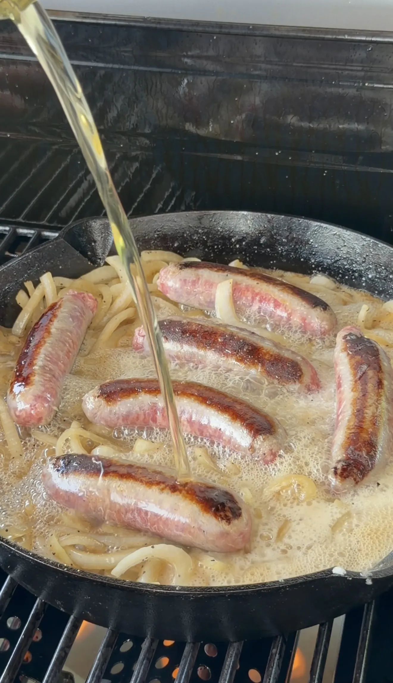 Bratwursts and sliced onions simmering in foamy beer in a cast iron skillet on a grill