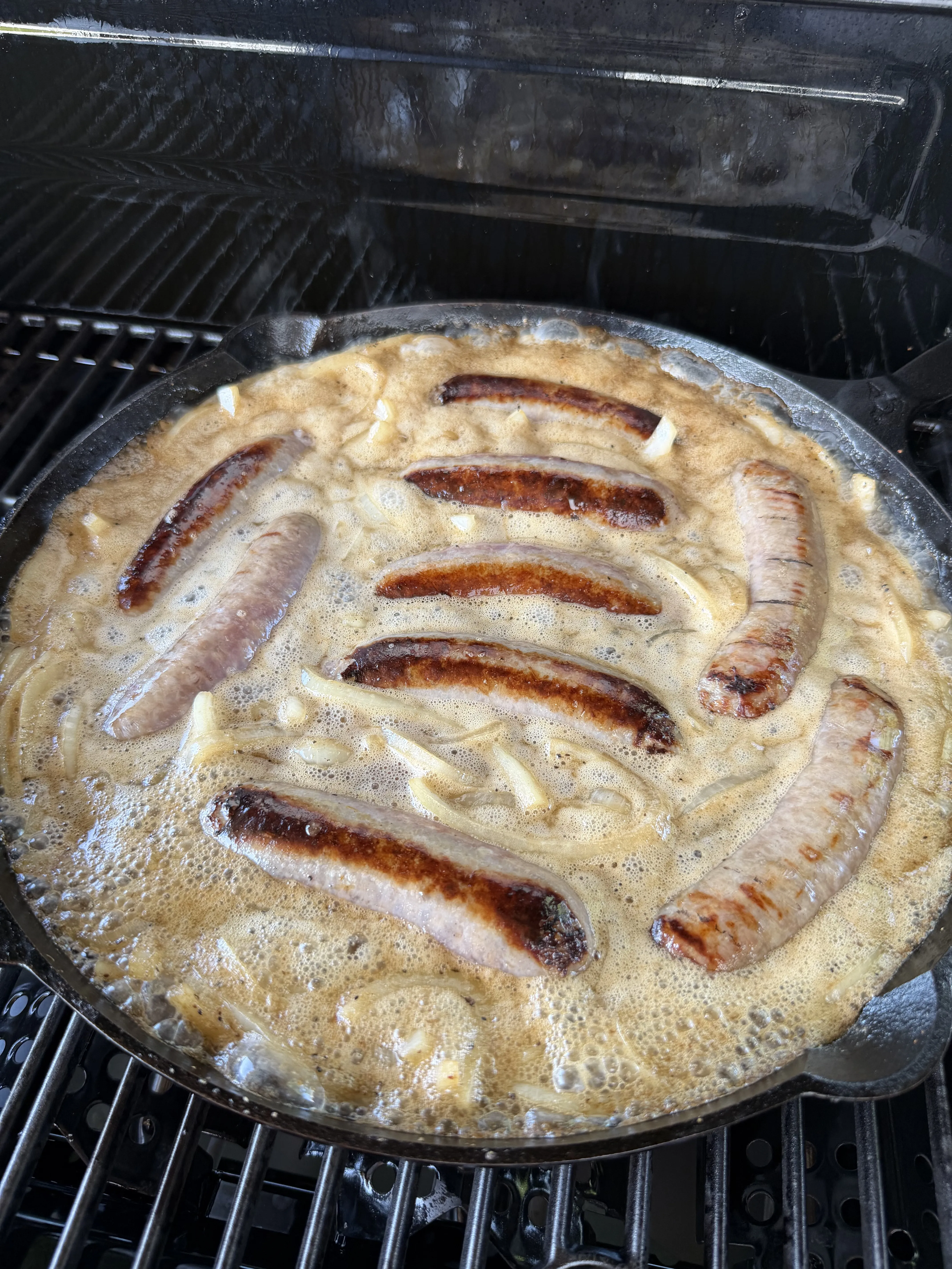 Brats simmering in beer with caramelized onions in a cast iron skillet on a grill