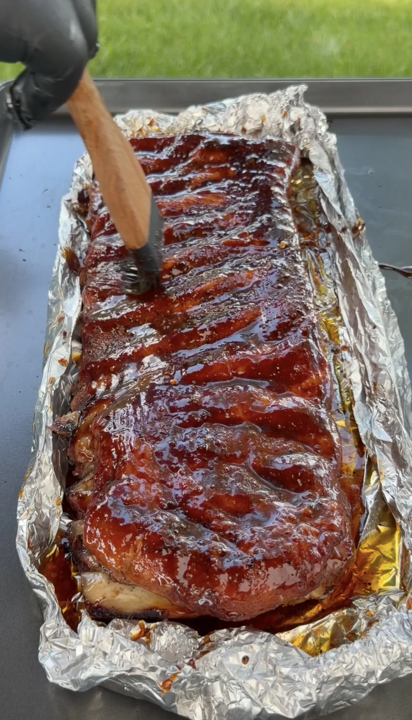 A rack of competition smoked spare ribs being brushed with glossy BBQ glaze on foil