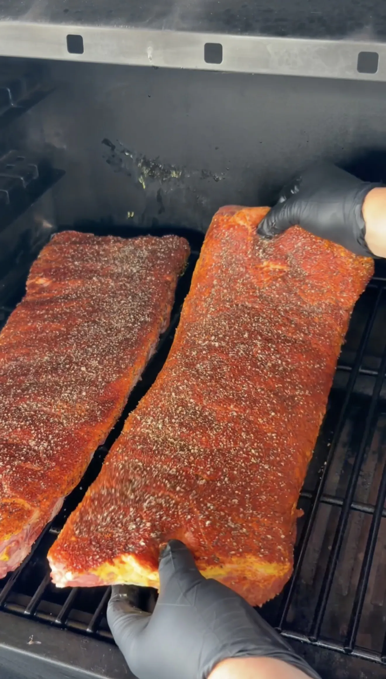 Two racks of competition spare ribs coated in rub being placed on smoker grates
