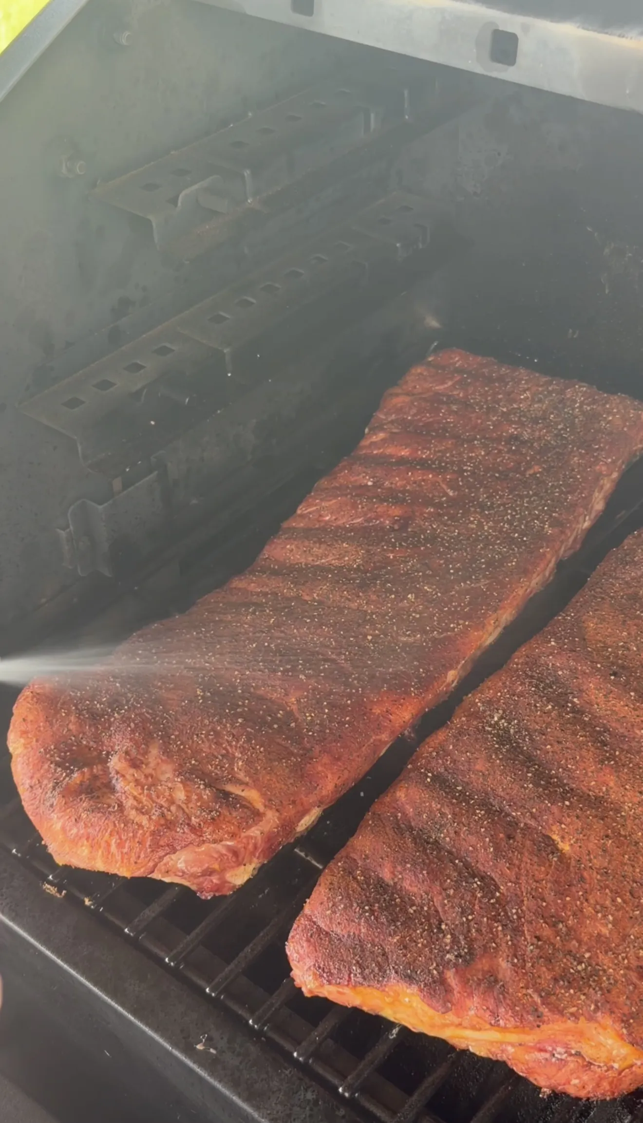 Two racks of competition style spare ribs building dark bark on grates inside a smoker