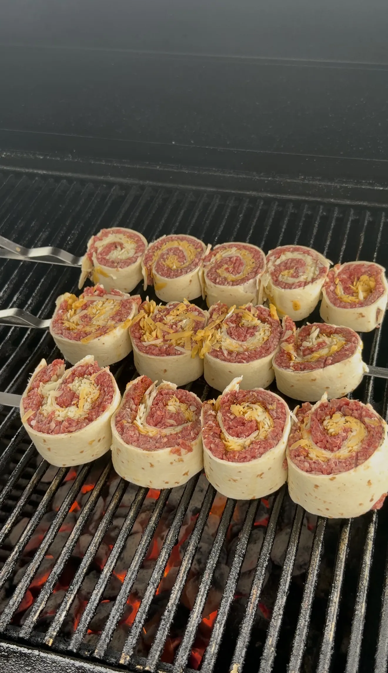 Beef tortilla pinwheels skewered on metal skewers over hot charcoal grill grates