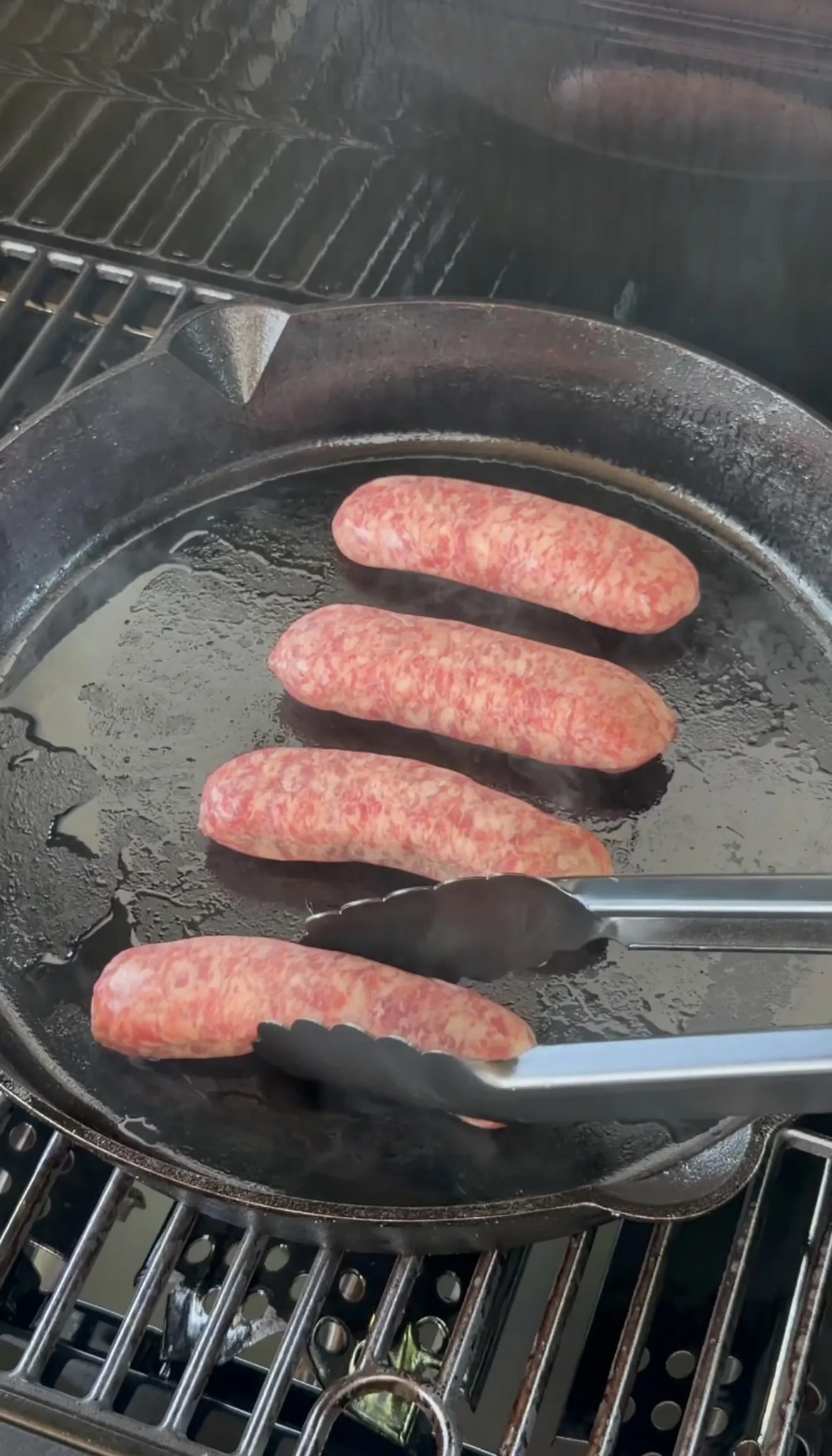 Raw bratwurst sausages searing in a cast iron skillet on a grill held with tongs