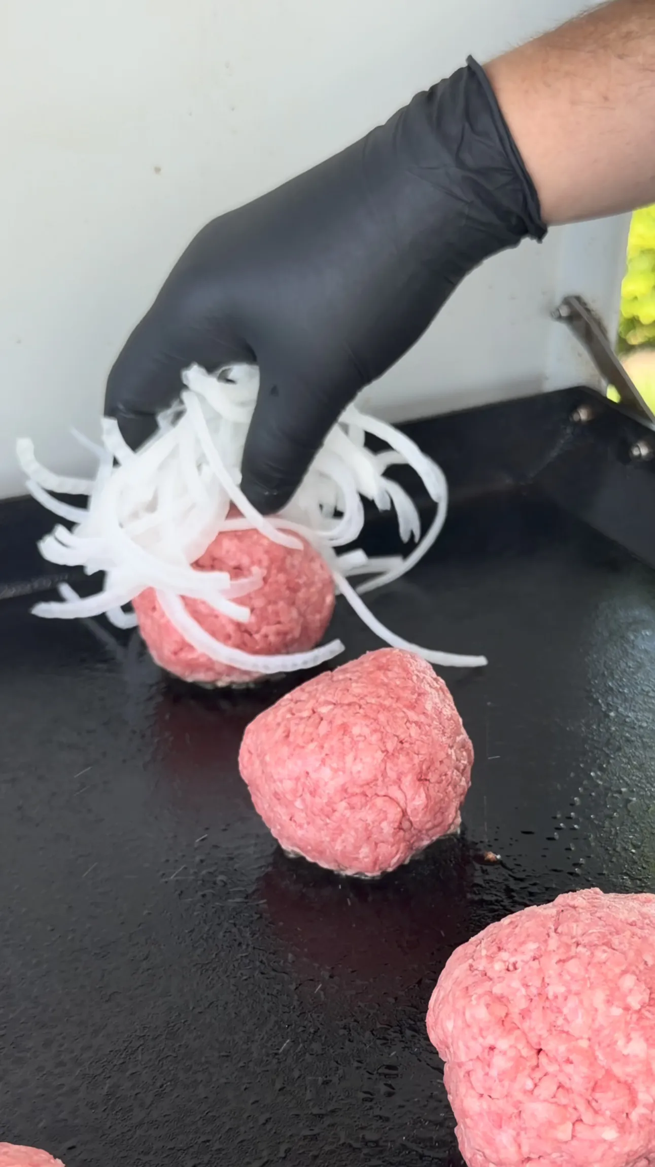 Raw beef balls topped with piles of shredded onions on the griddle before smashing