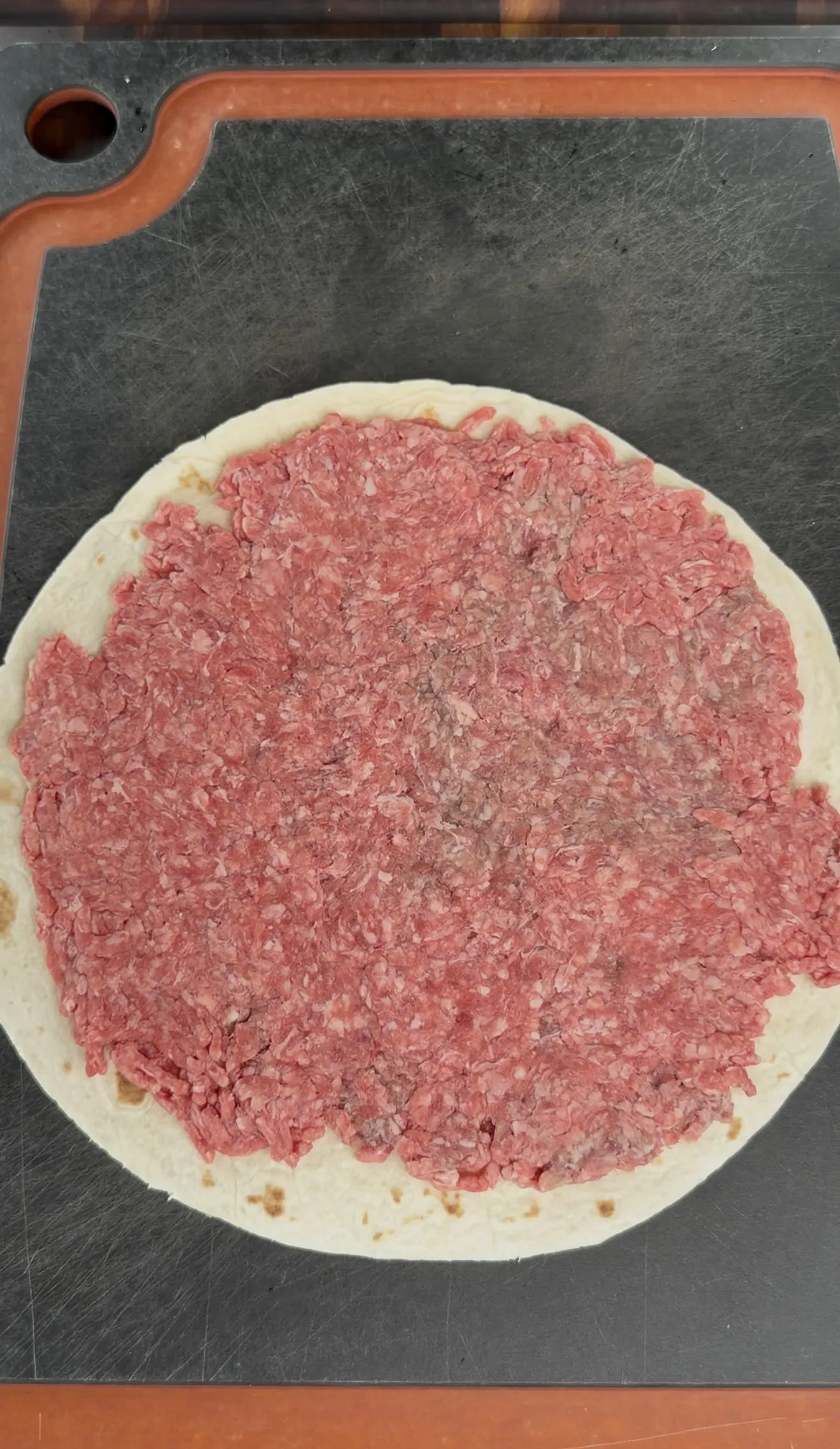 Raw seasoned ground beef spread evenly over a flour tortilla for pinwheel prep