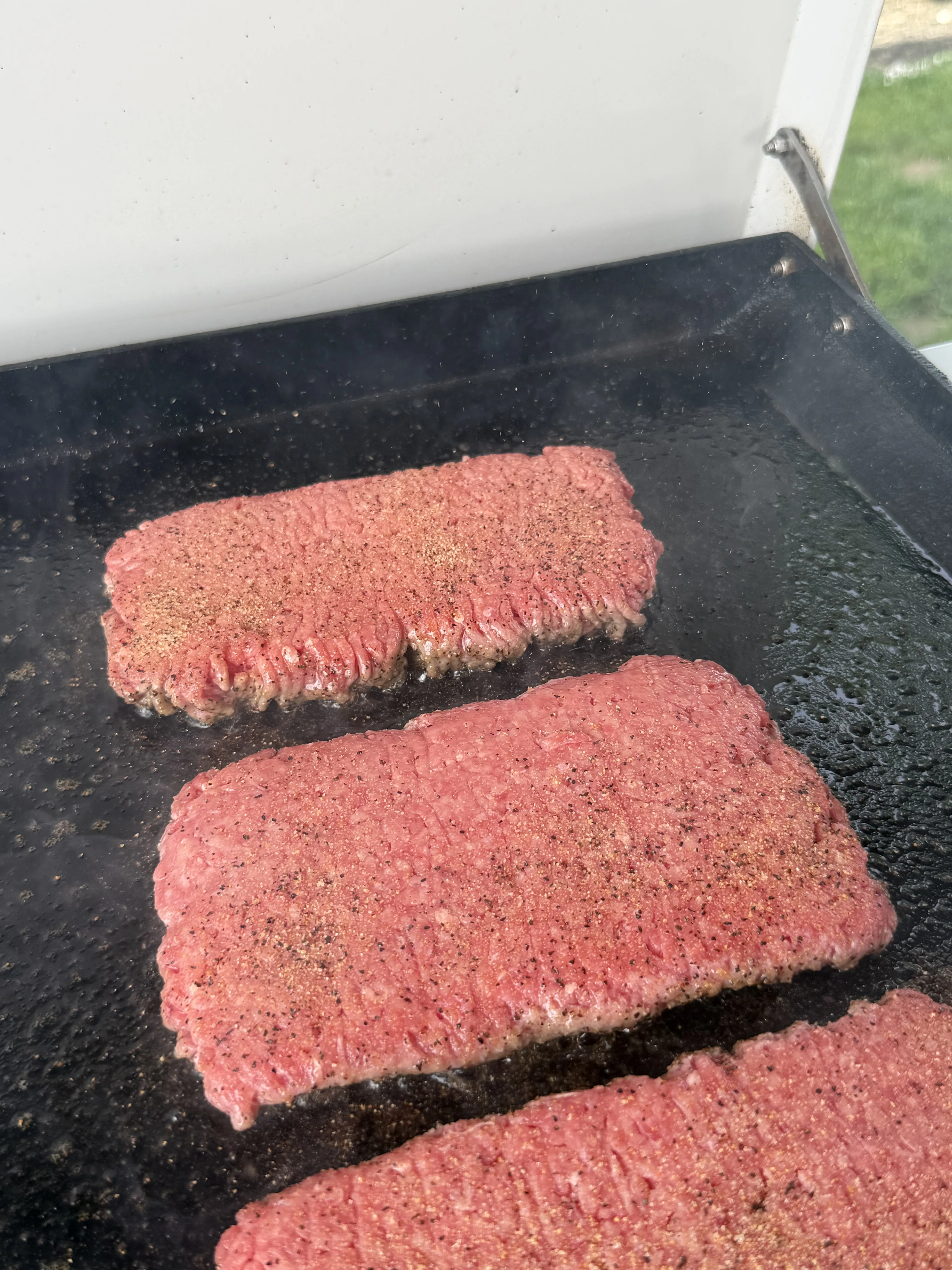 Raw seasoned beef patties cooking on a black griddle for bacon jam patty melts