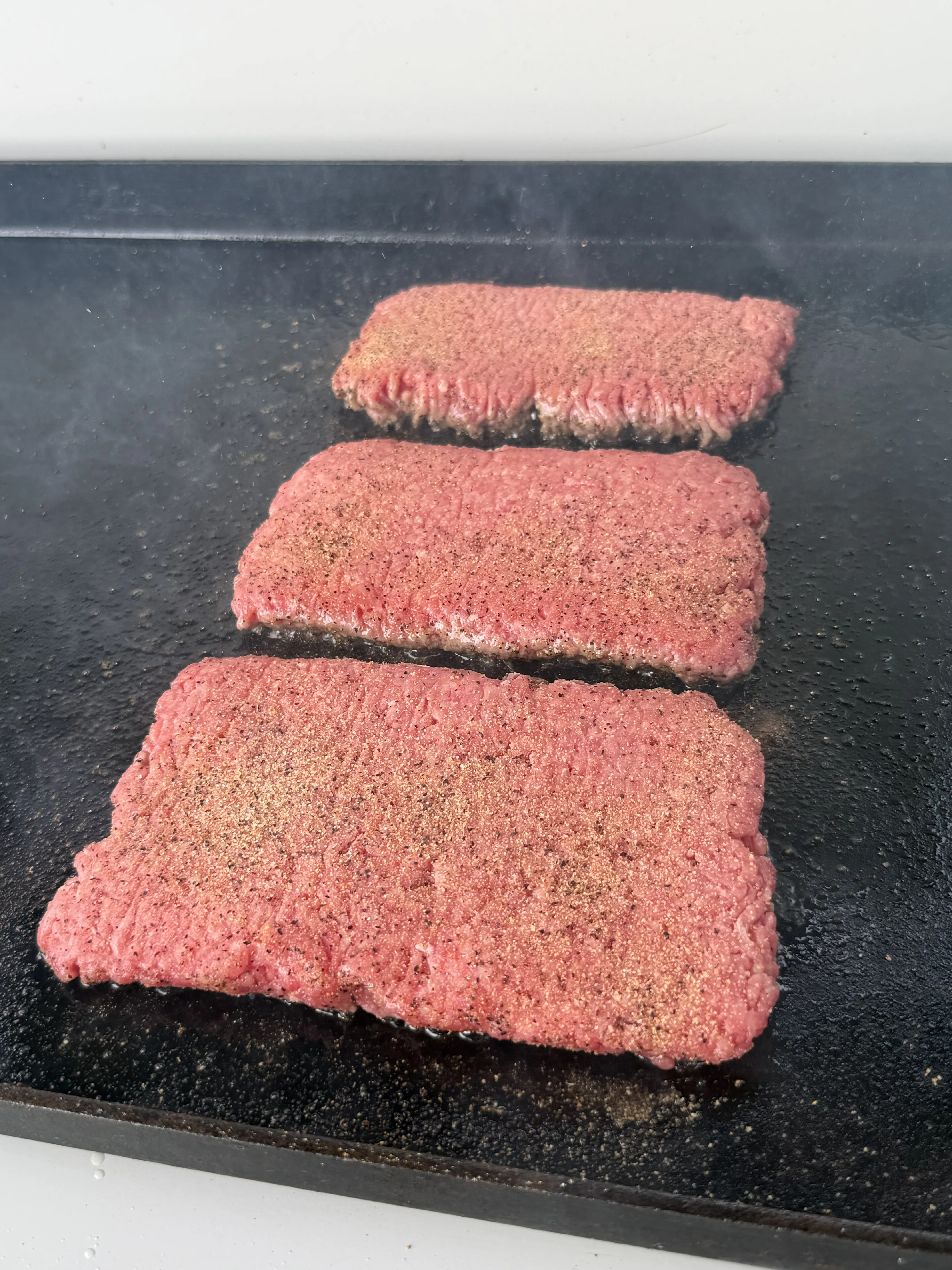 Three seasoned beef patties cooking on a griddle for bacon jam patty melts