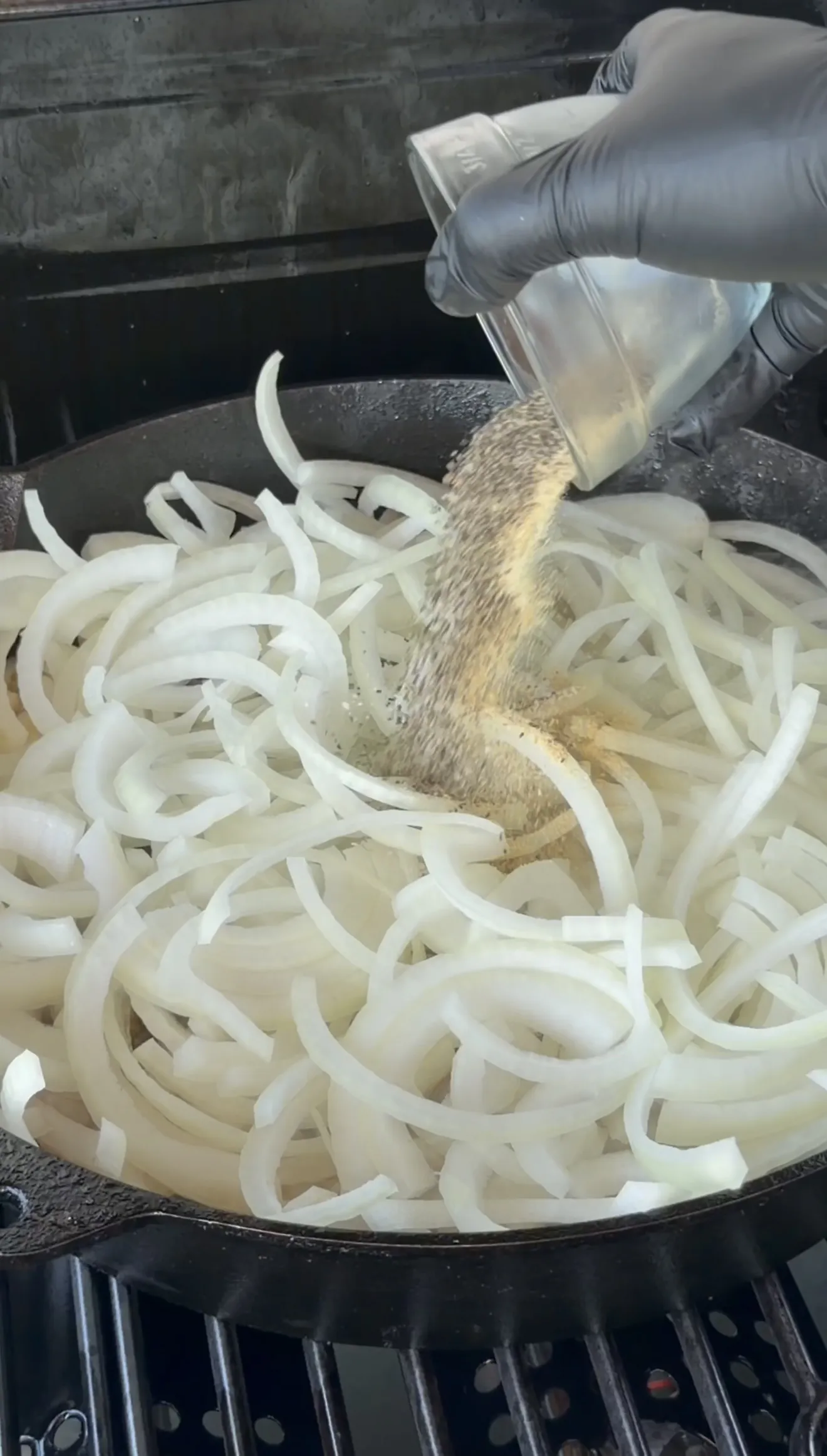Gloved hand seasoning thinly sliced onions in a cast iron skillet for beer brats