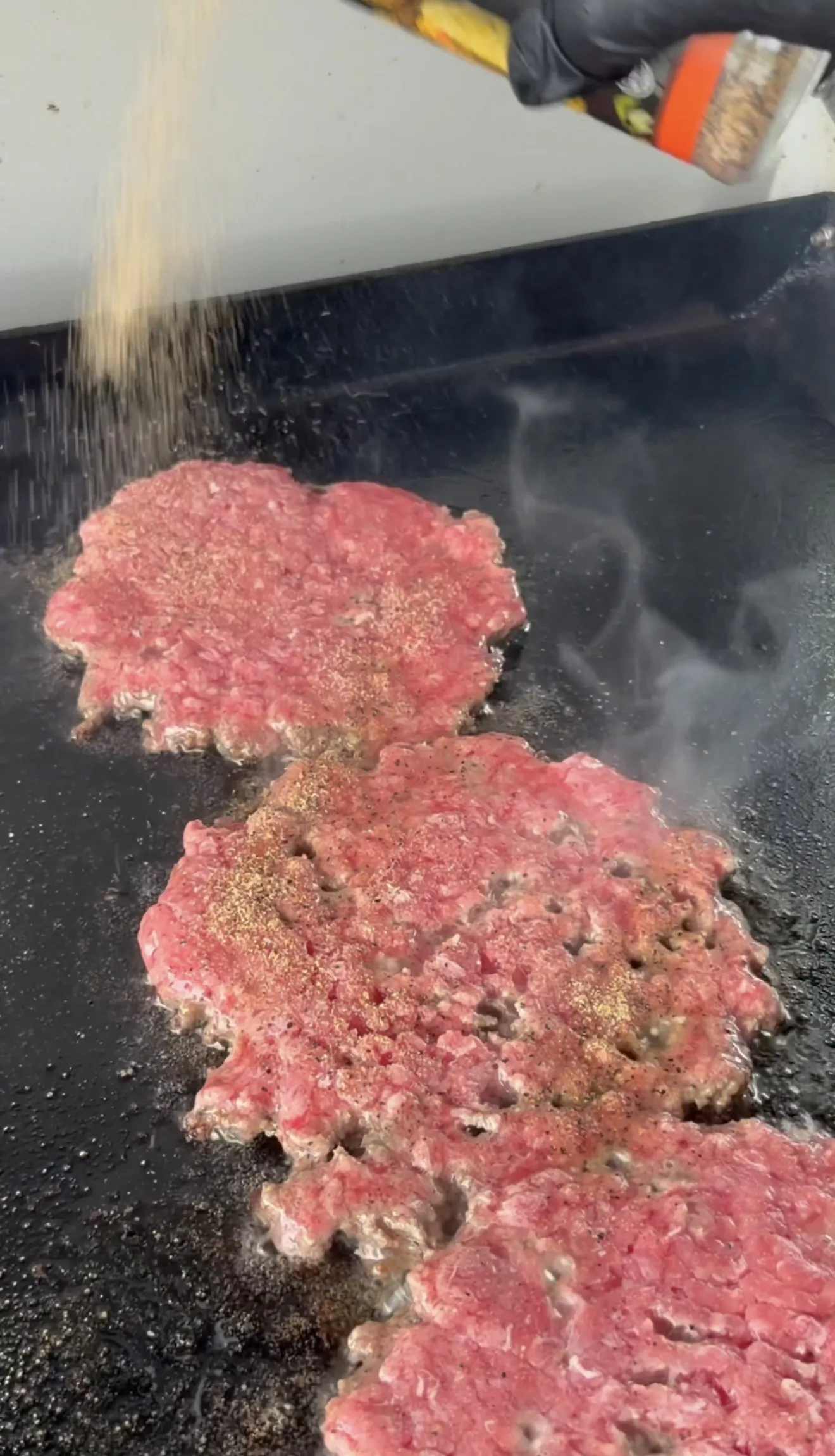 Smash burger patties being seasoned on the griddle before flipping
