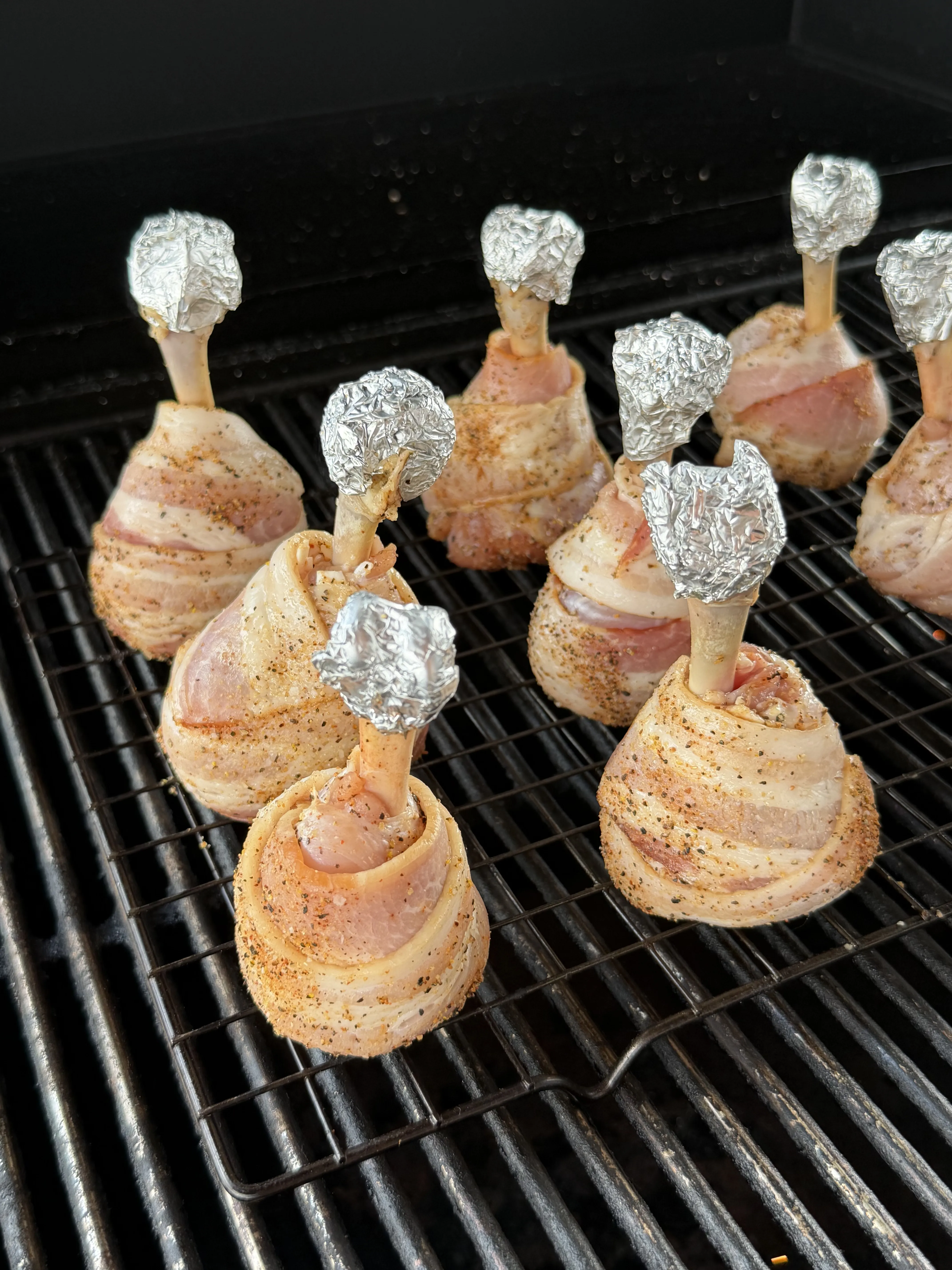 Seasoned bacon wrapped chicken lollipops standing upright on smoker grates with foil-tipped bones