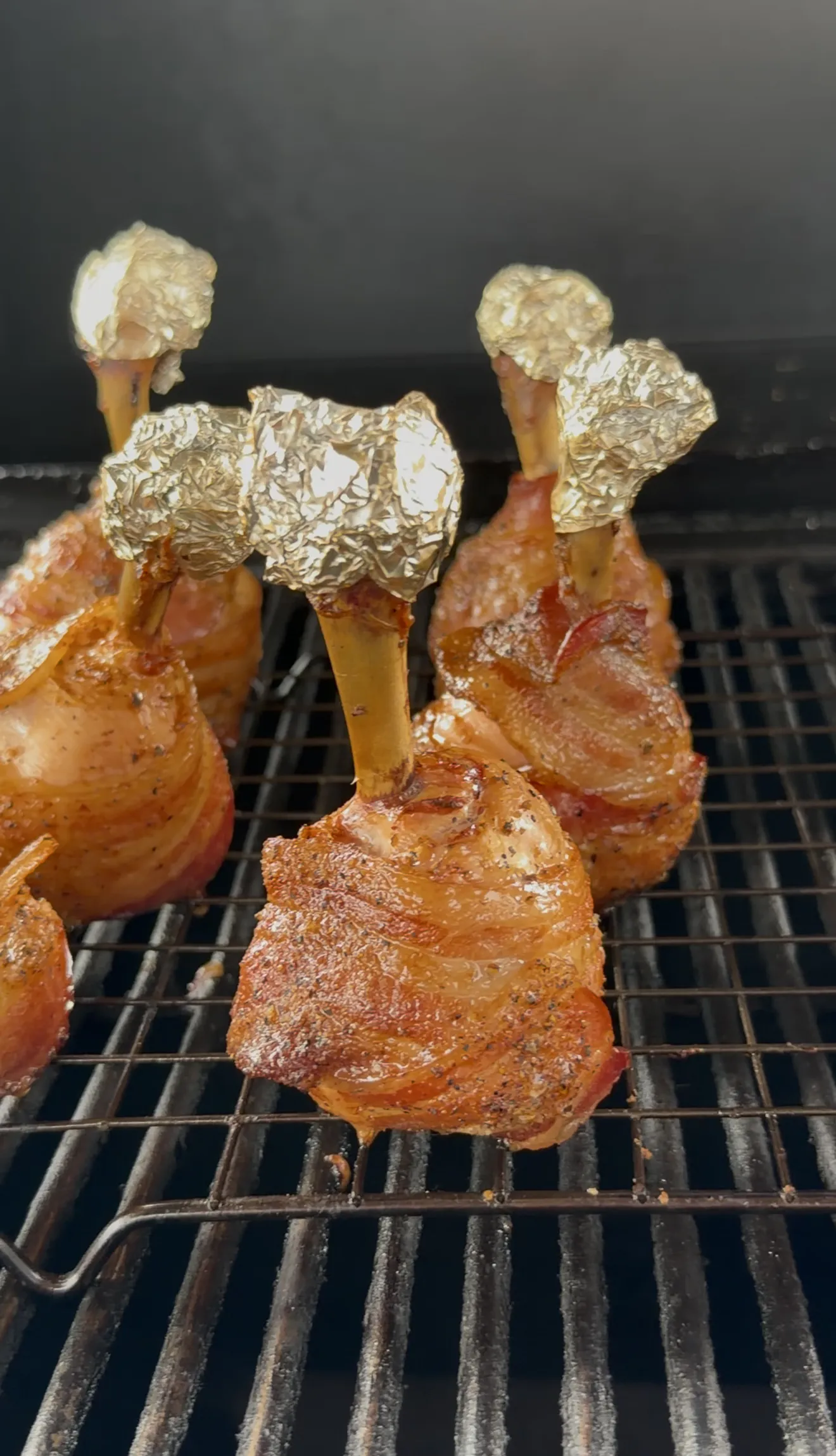 Molasses candied bacon wrapped chicken lollipops with foil-tipped bones smoking on the grate