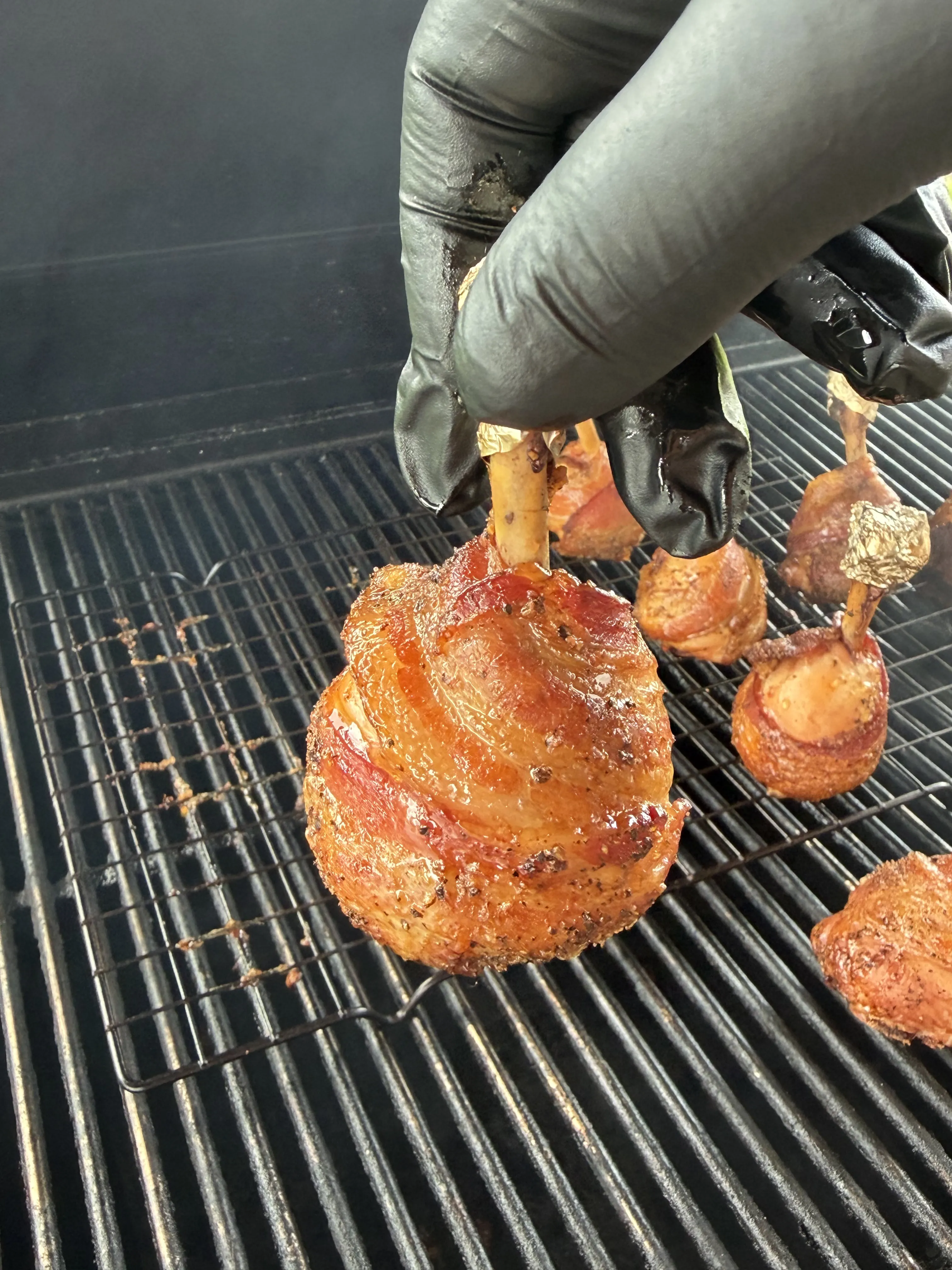 Molasses candied bacon wrapped chicken lollipops on smoker grill grates held by a gloved hand