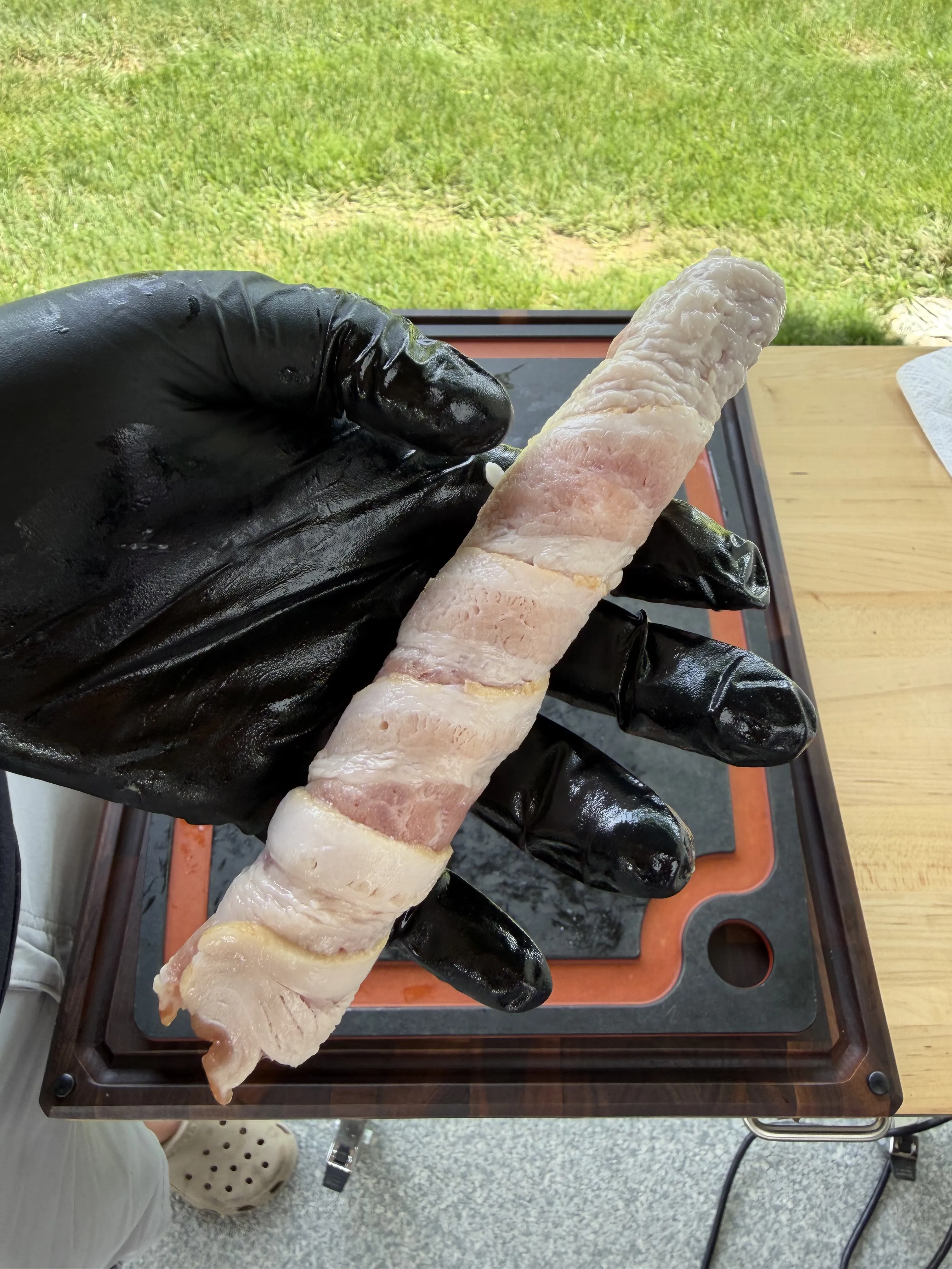 A raw bacon wrapped hot dog held over a cutting board, ready for the smoker