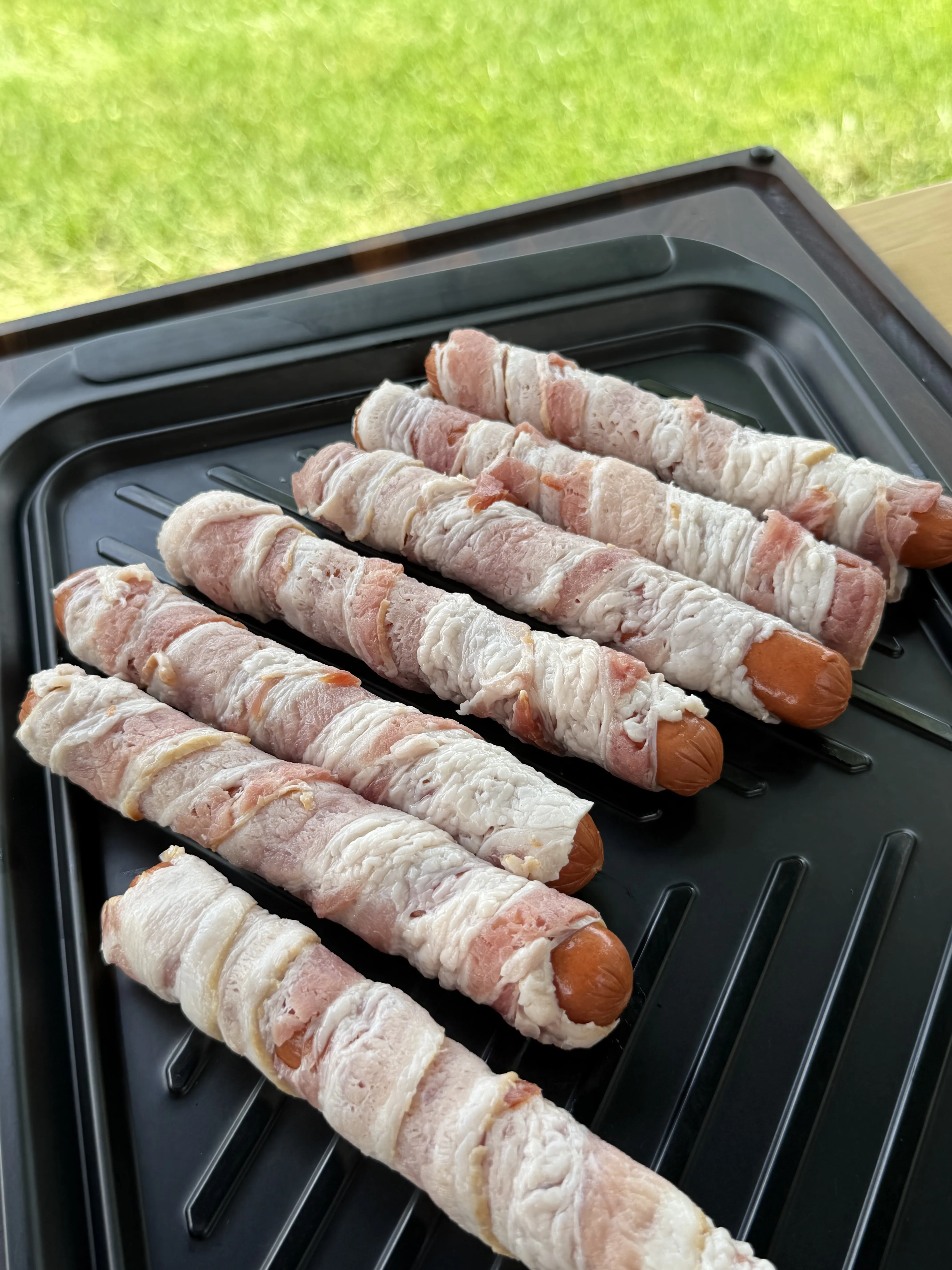 Raw bacon wrapped hot dogs arranged on a grill pan, ready to cook