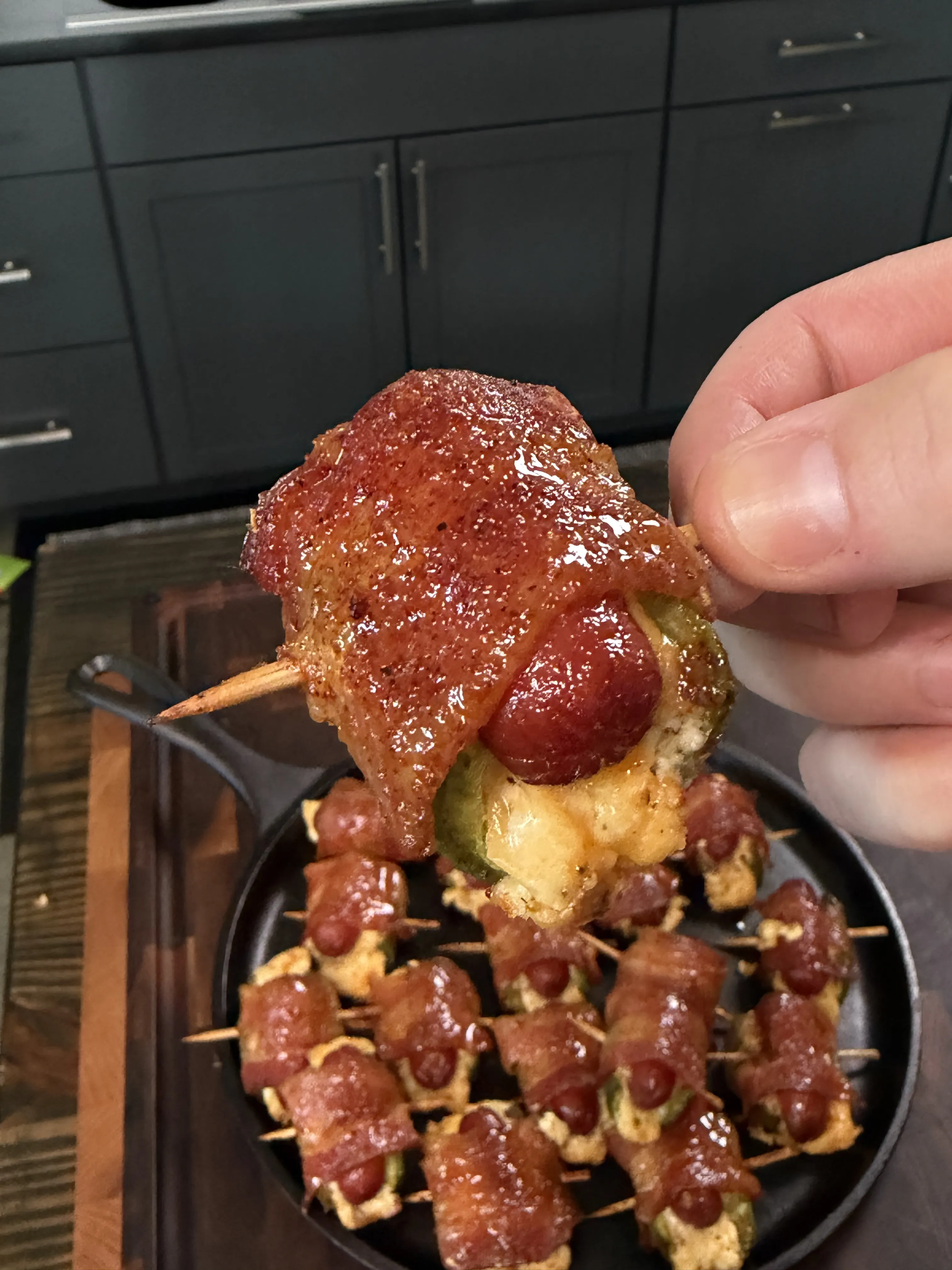 Hand holding a glazed bacon wrapped jalapeño smokie bomb over a cast iron pan of more