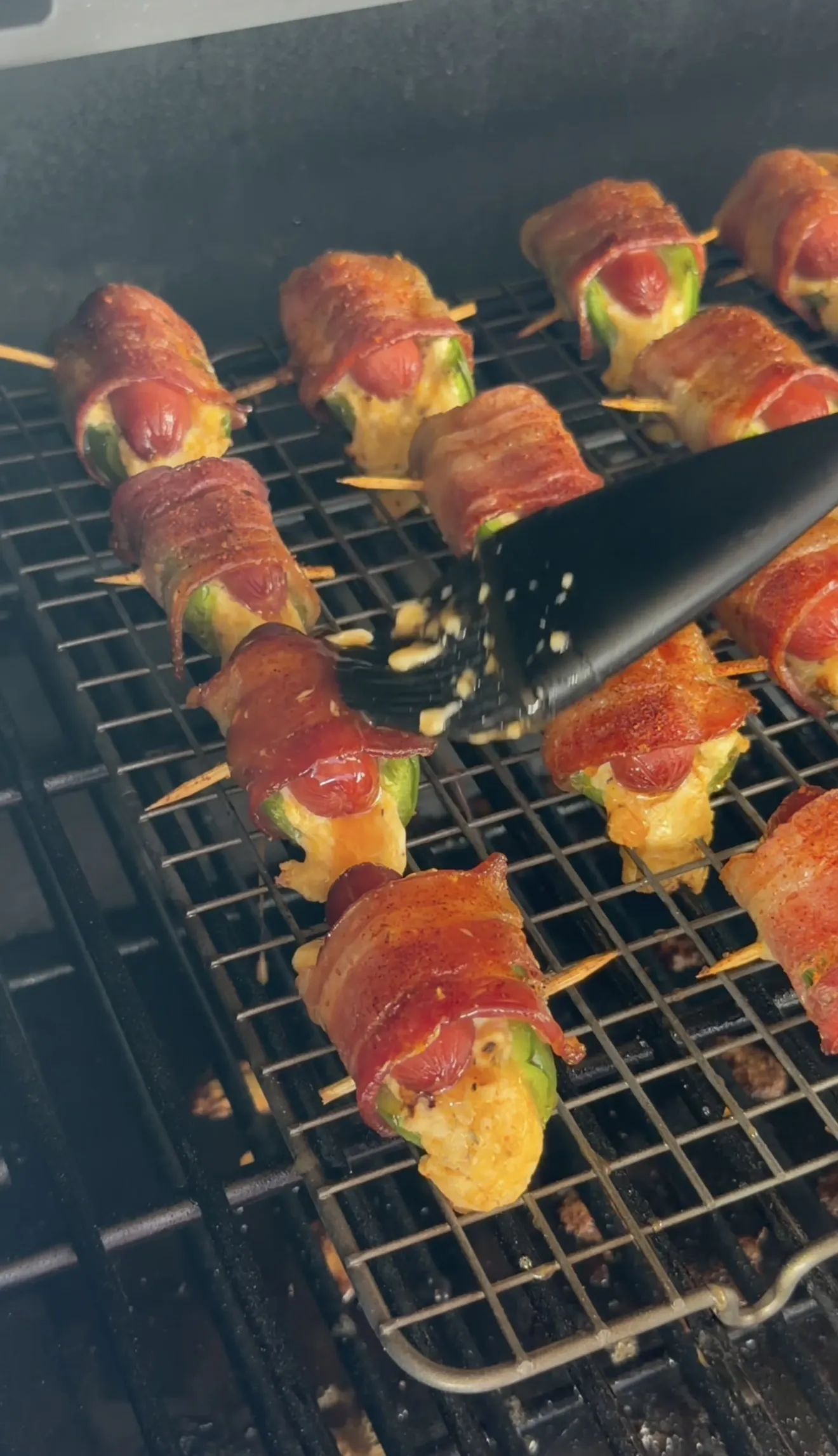 Bacon wrapped jalapeño smokie bombs with melted cheese on a smoker rack being basted with sauce