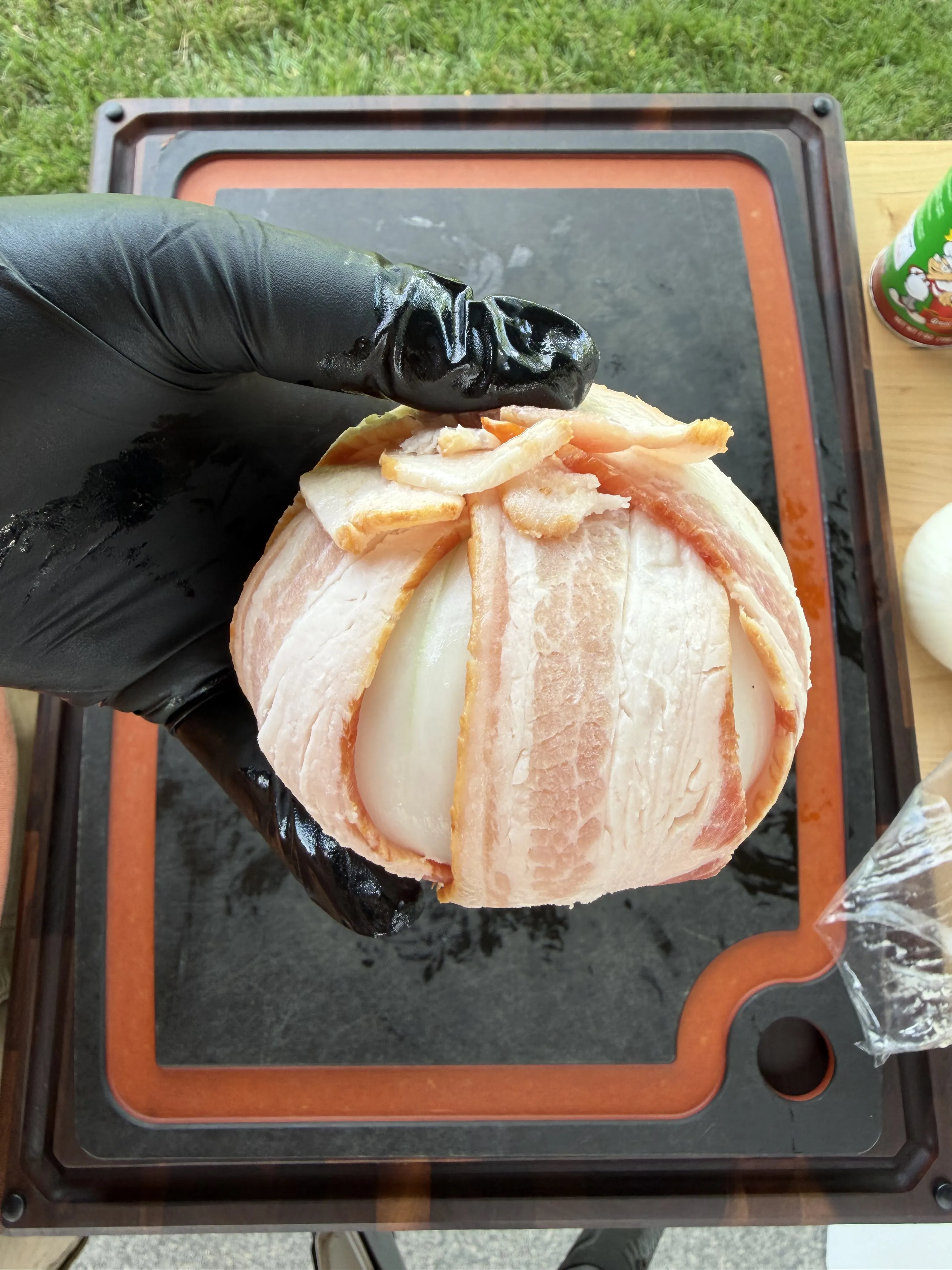 Stuffed onion being wrapped in overlapping bacon strips over a black cutting board