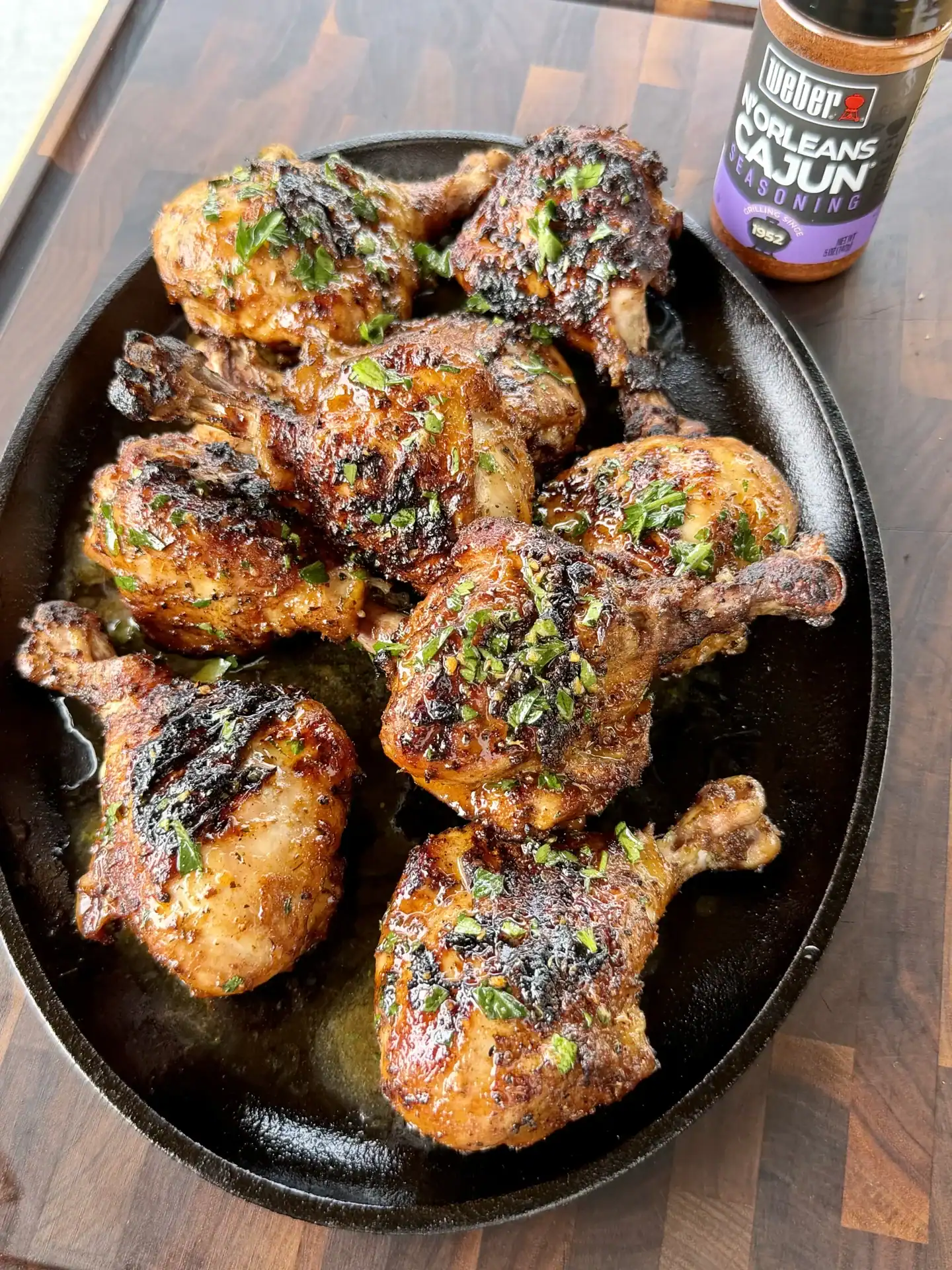 Juicy grilled Cajun chicken drumsticks on a cast iron platter, garnished with fresh herbs, with Weber N'Orleans Cajun seasoning nearby.