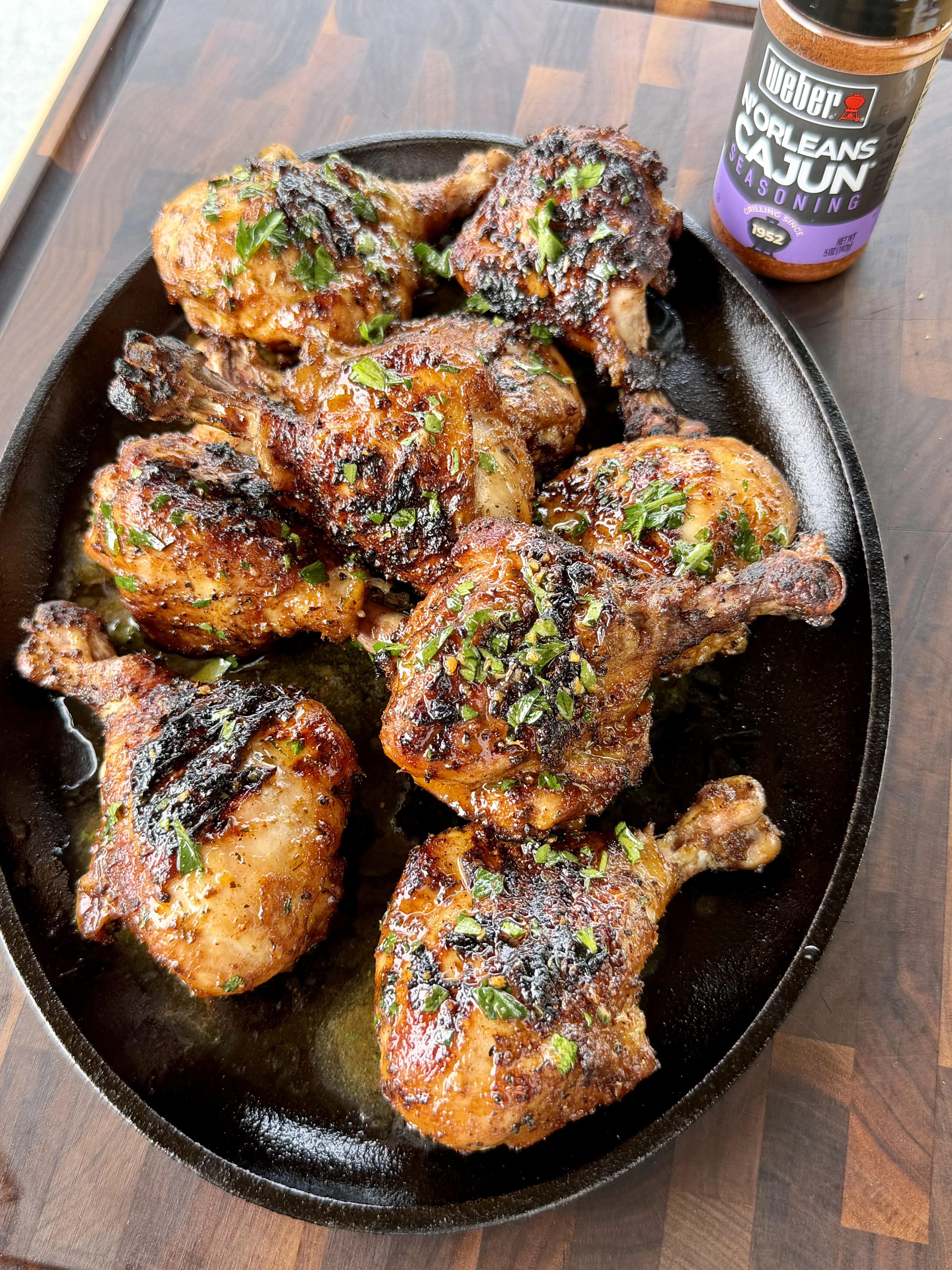 Juicy grilled Cajun chicken drumsticks on a cast iron platter, garnished with fresh herbs, with Weber N'Orleans Cajun seasoning nearby