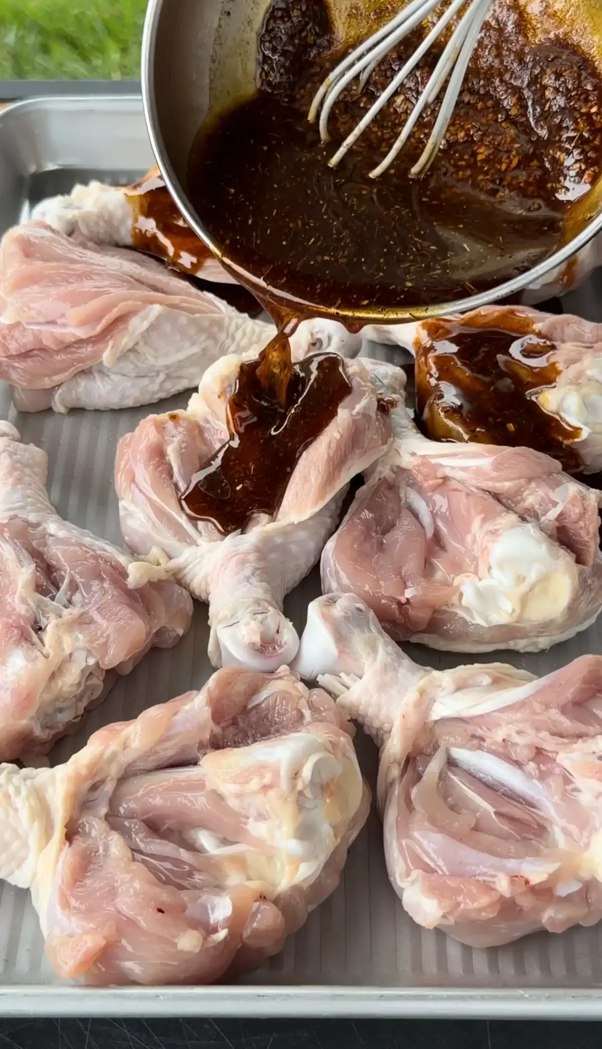 Dark Cajun marinade being poured from a bowl over raw chicken drumsticks on a metal baking sheet.