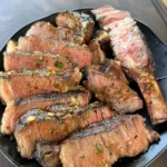 Grilled beef ribs sliced and piled on a black plate, coated in chili parmesan compound butter with herbs and garlic