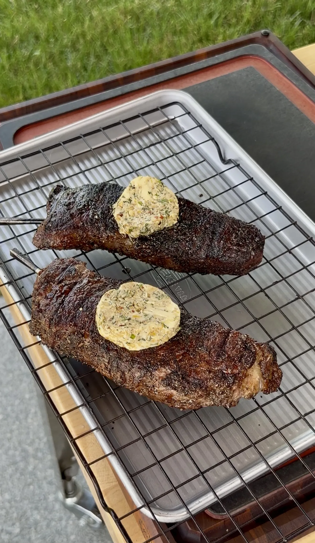 Two seared steaks resting on a wire rack topped with melting butter discs