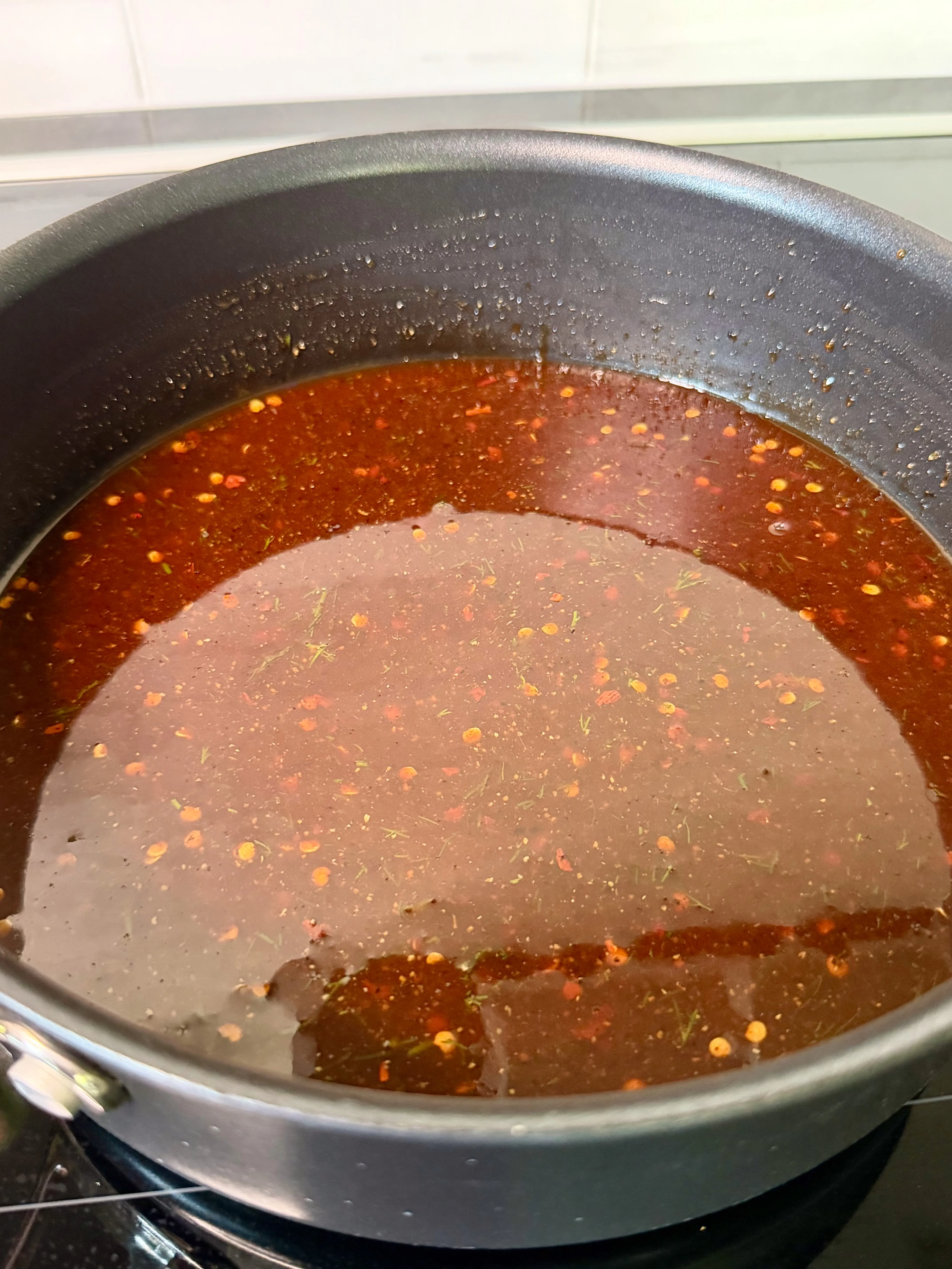 Deep red dill pickle hot honey bbq sauce with chili flakes and herbs simmering in a dark skillet