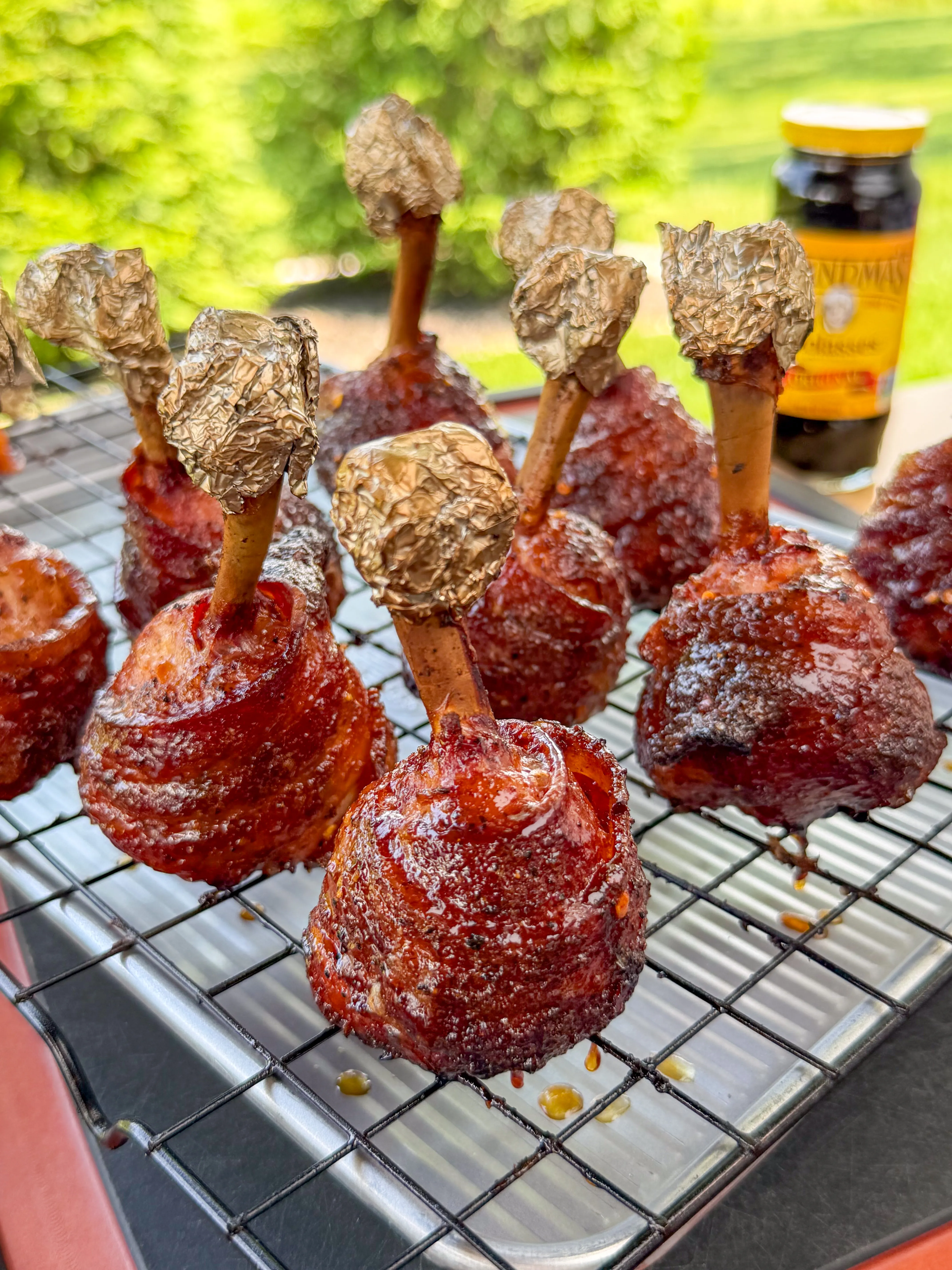 Glazed bacon-wrapped chicken lollipops with foil-tipped bones on a wire rack, coated in molasses BBQ glaze