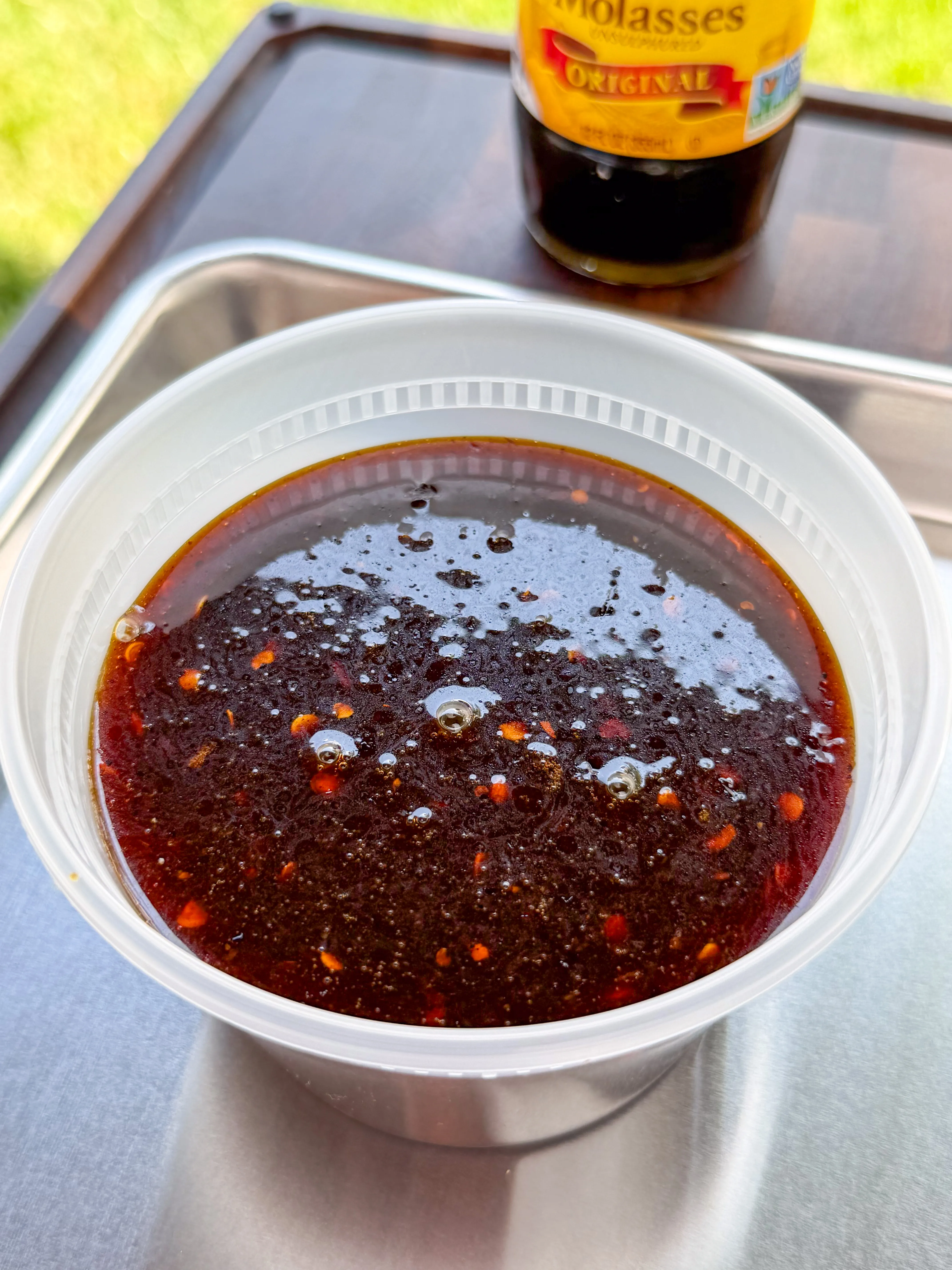Dark molasses BBQ glaze with red pepper flakes in a plastic bowl, with a bottle of original molasses in the background