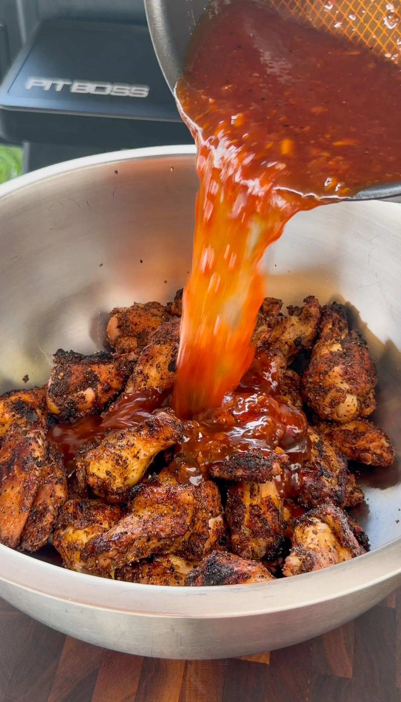 Orange gochujang BBQ sauce poured over smoked wings in a steel mixing bowl for tossing