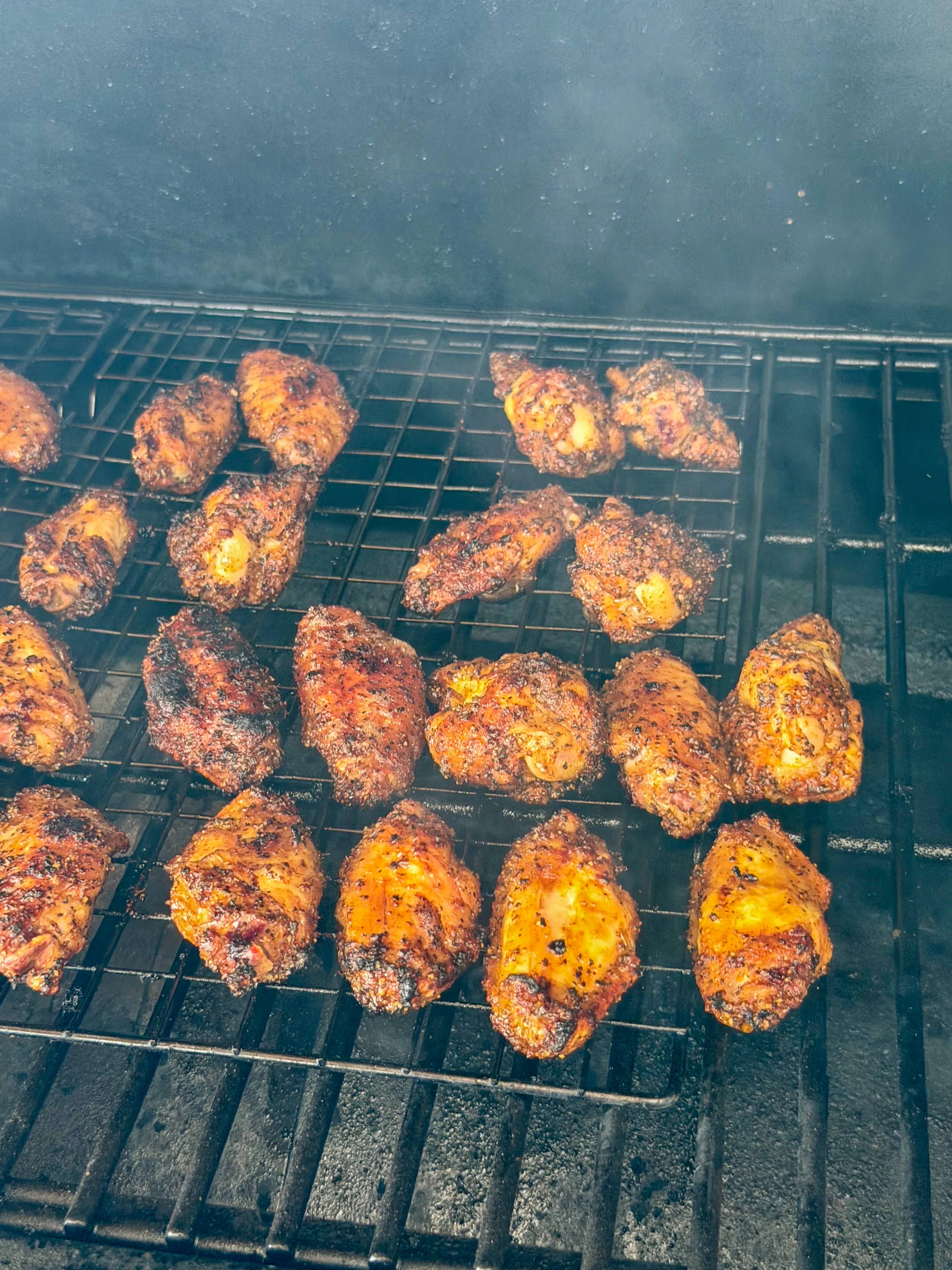 Smoky wings cooking on a smoker grill with visible smoke rolling through