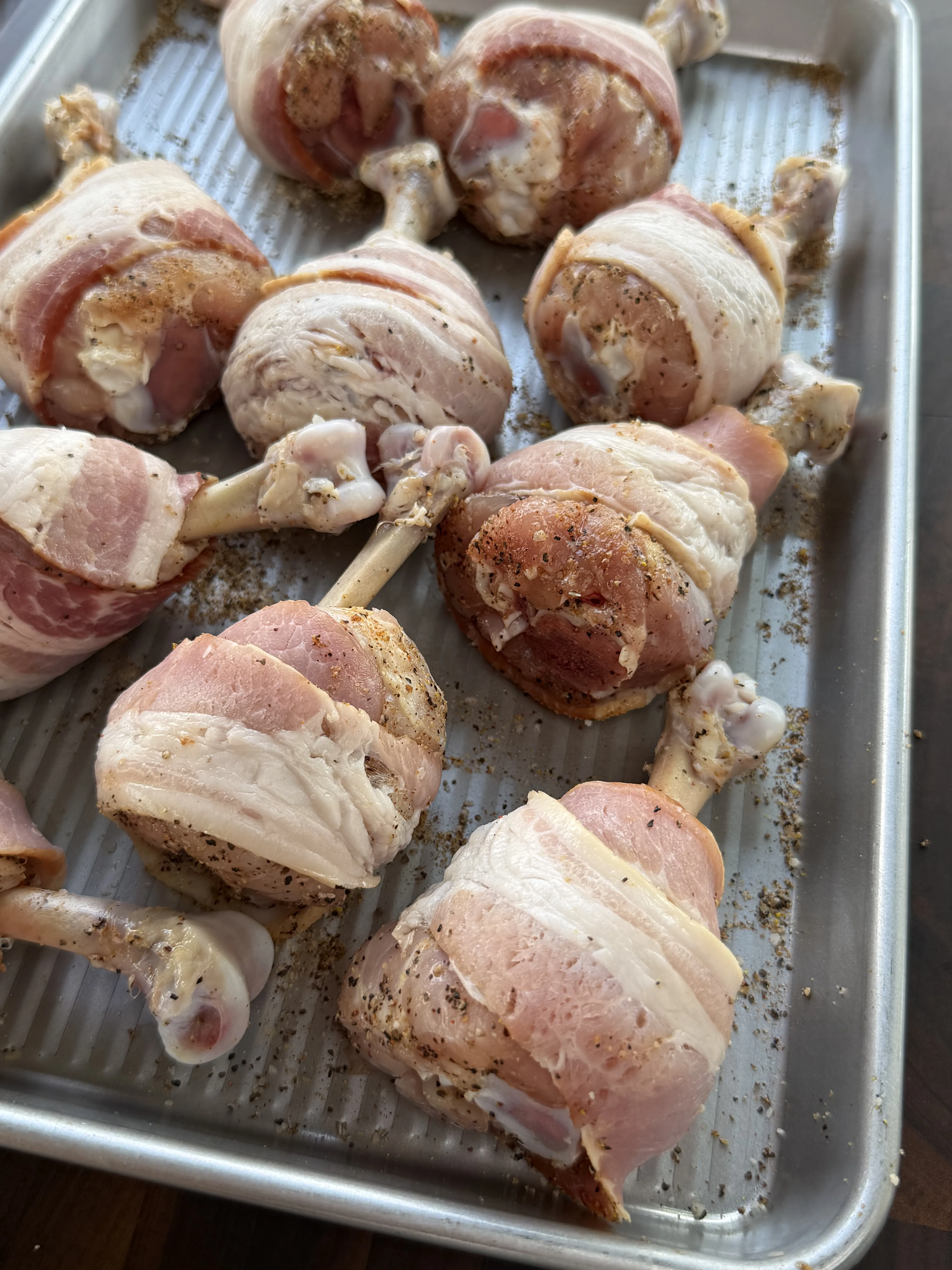 Raw bacon wrapped chicken lollipops seasoned and arranged on a baking sheet, ready to smoke