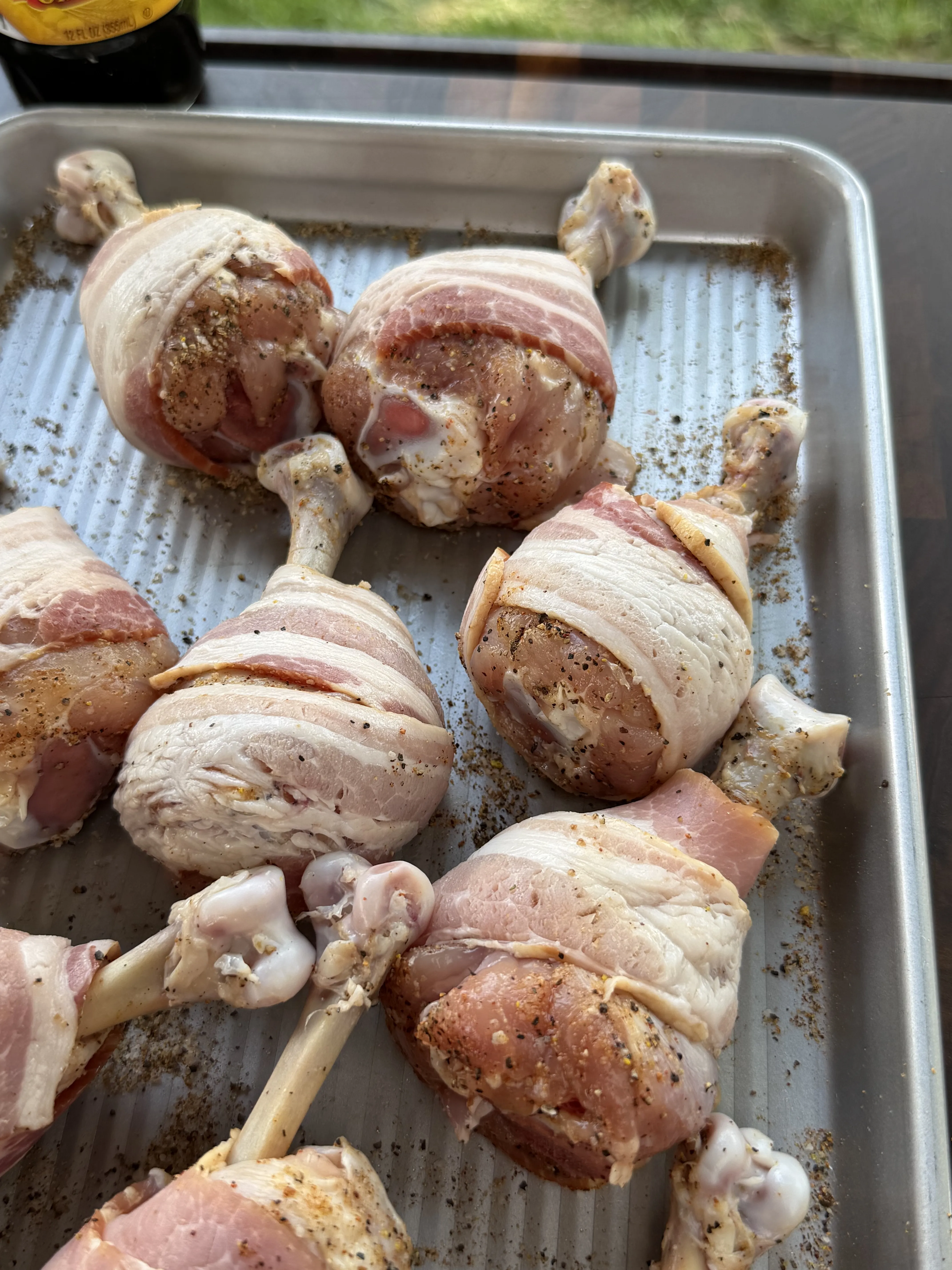Raw bacon wrapped chicken drumstick lollipops seasoned with spices on a metal baking sheet