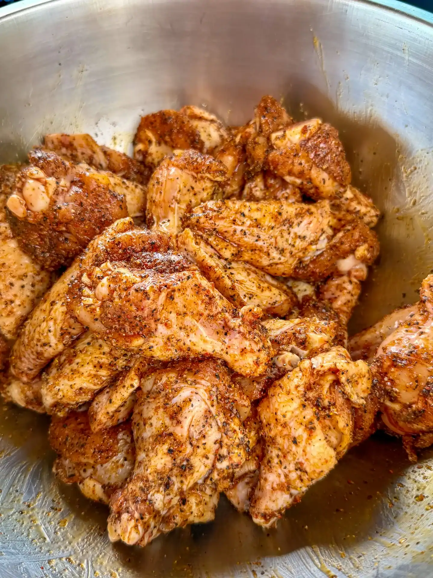 Raw chicken wings coated in smoky dry rub seasoning in a stainless steel mixing bowl.