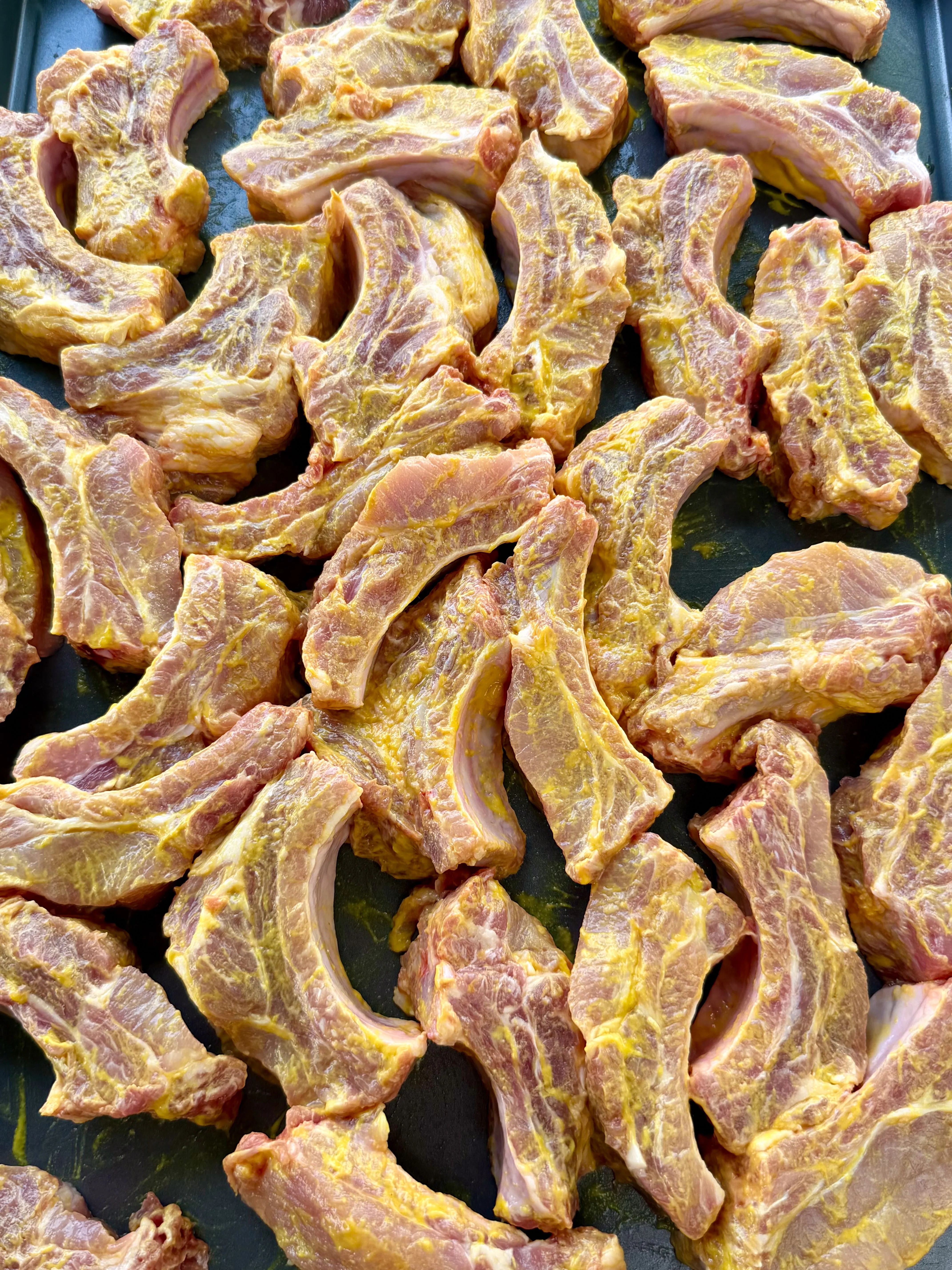 Raw party ribs coated in yellow mustard binder and spice rub on a dark baking sheet, ready to smoke