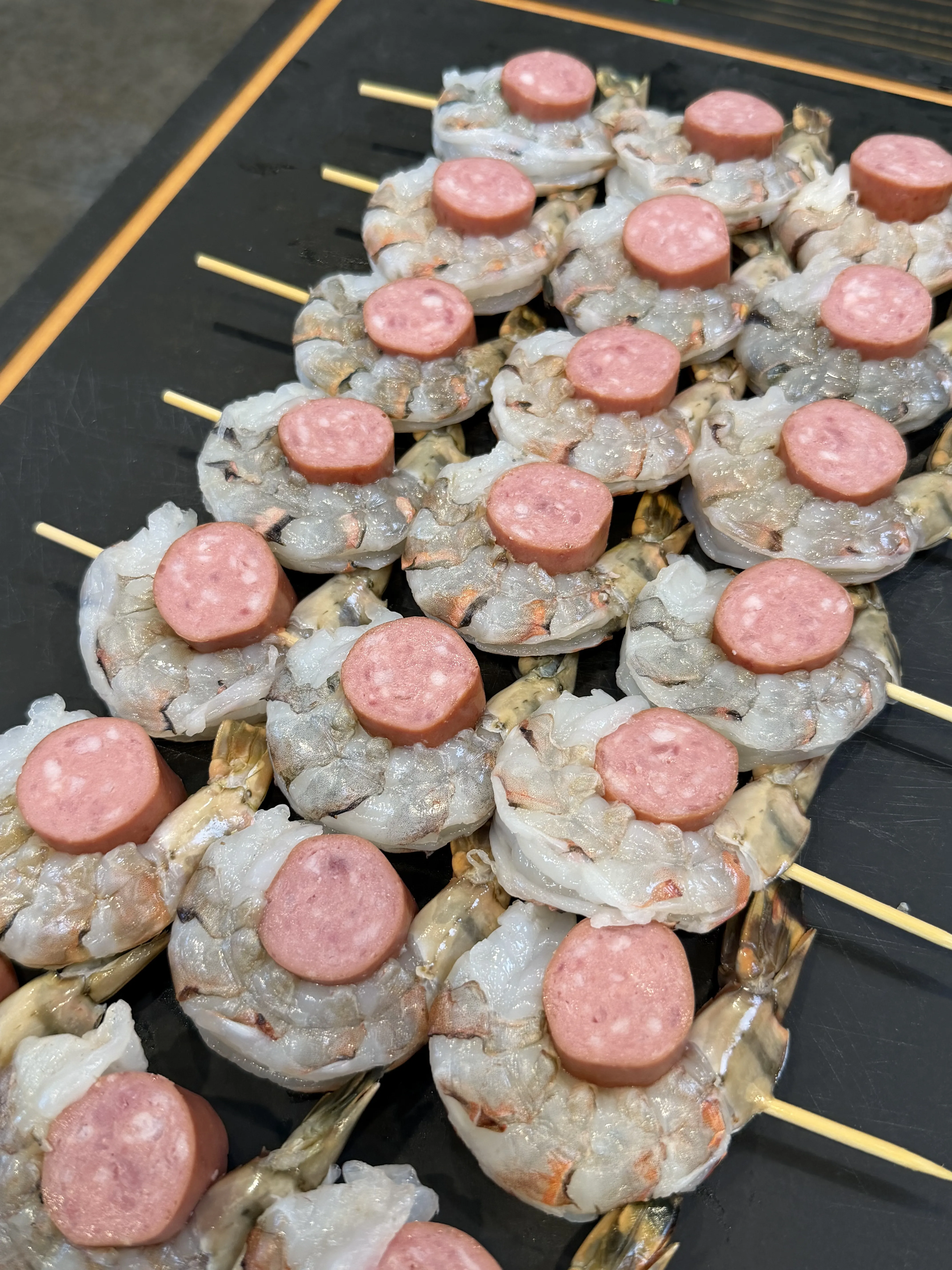 Raw shrimp and sausage rounds threaded on bamboo skewers arranged on a dark tray before smoking