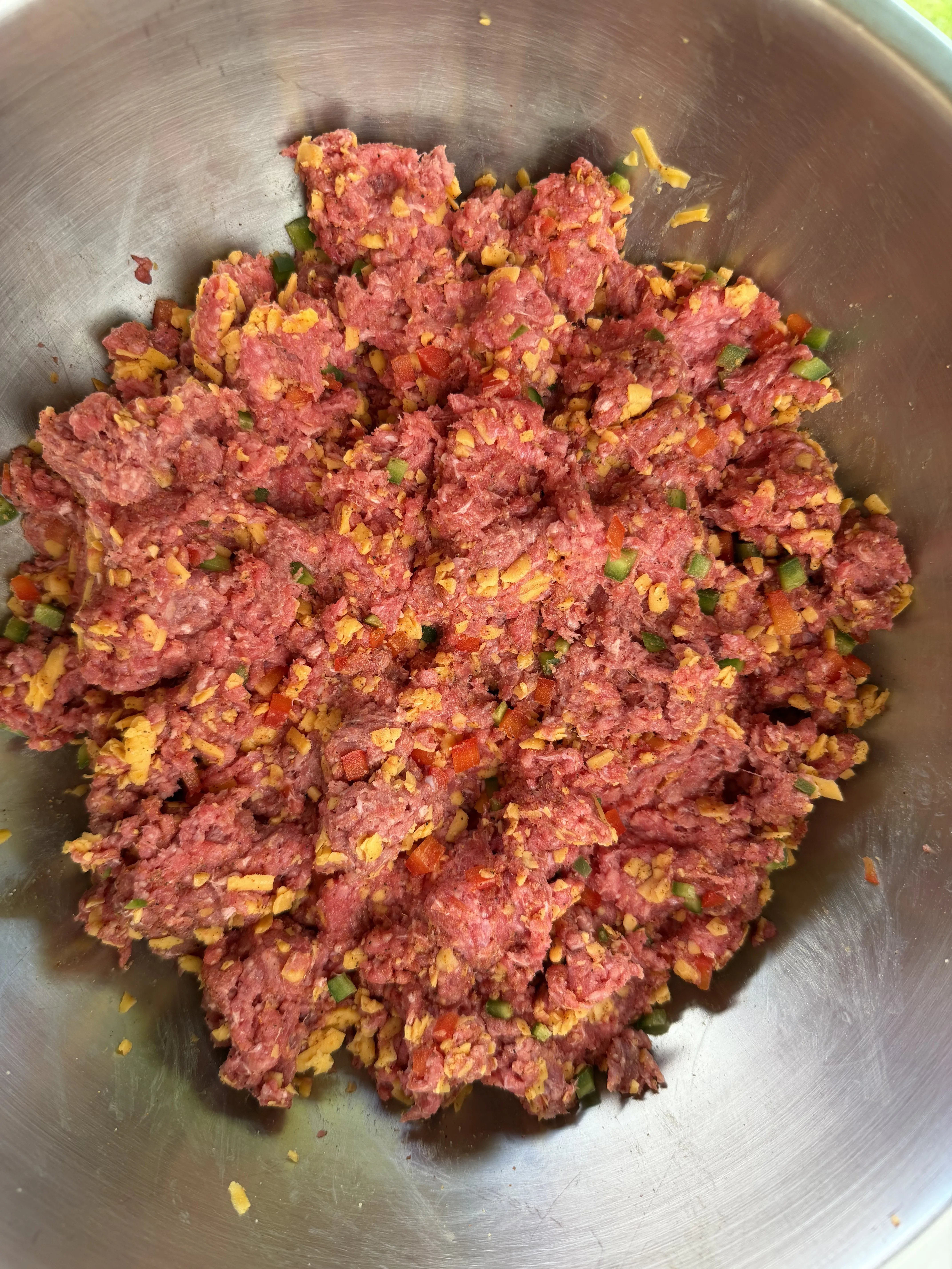 Ground beef mixed with shredded cheese, diced peppers, and seasoning in a stainless bowl