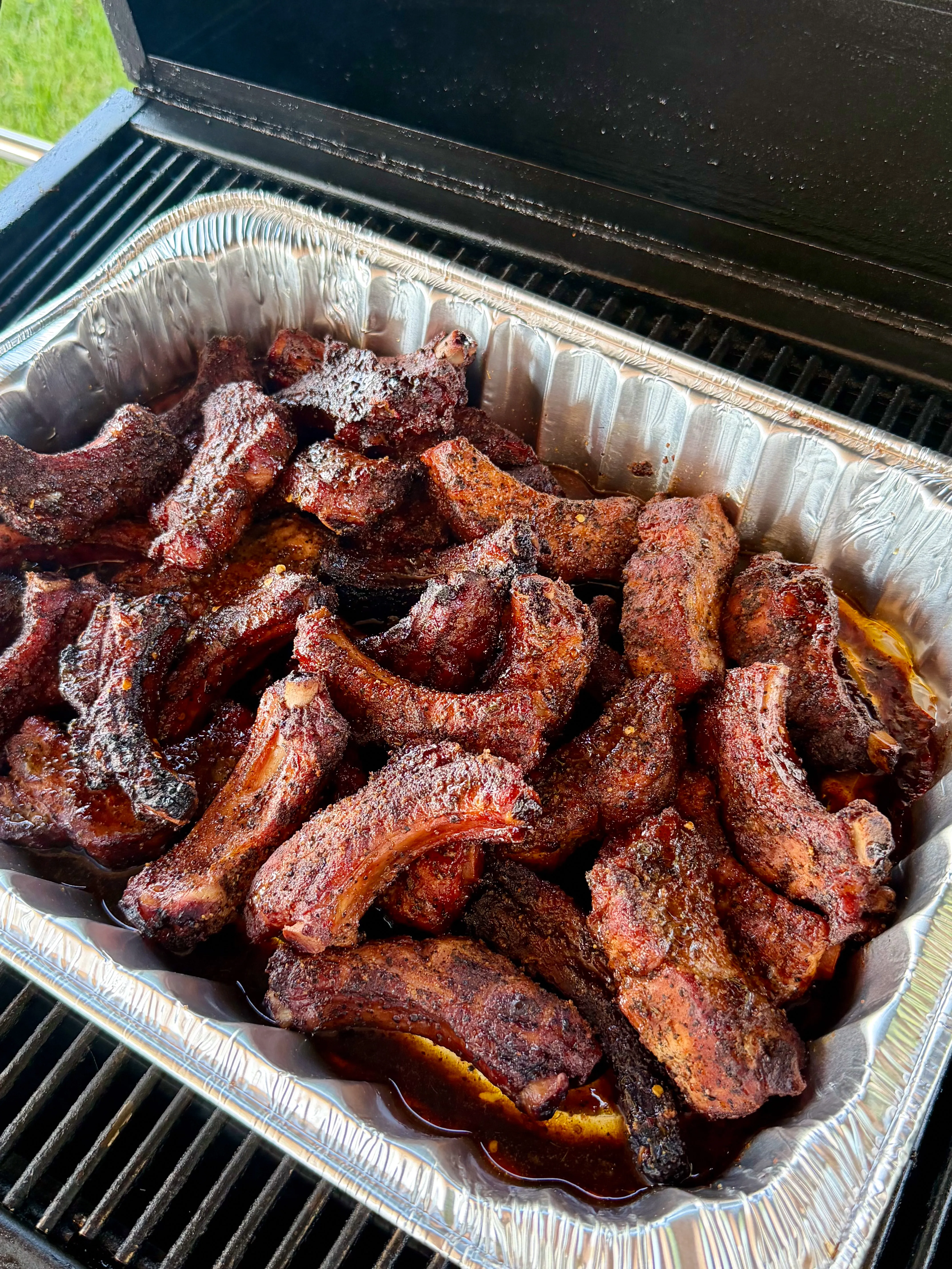 Smoky party ribs piled in a foil pan on a grill, coated in dark glaze