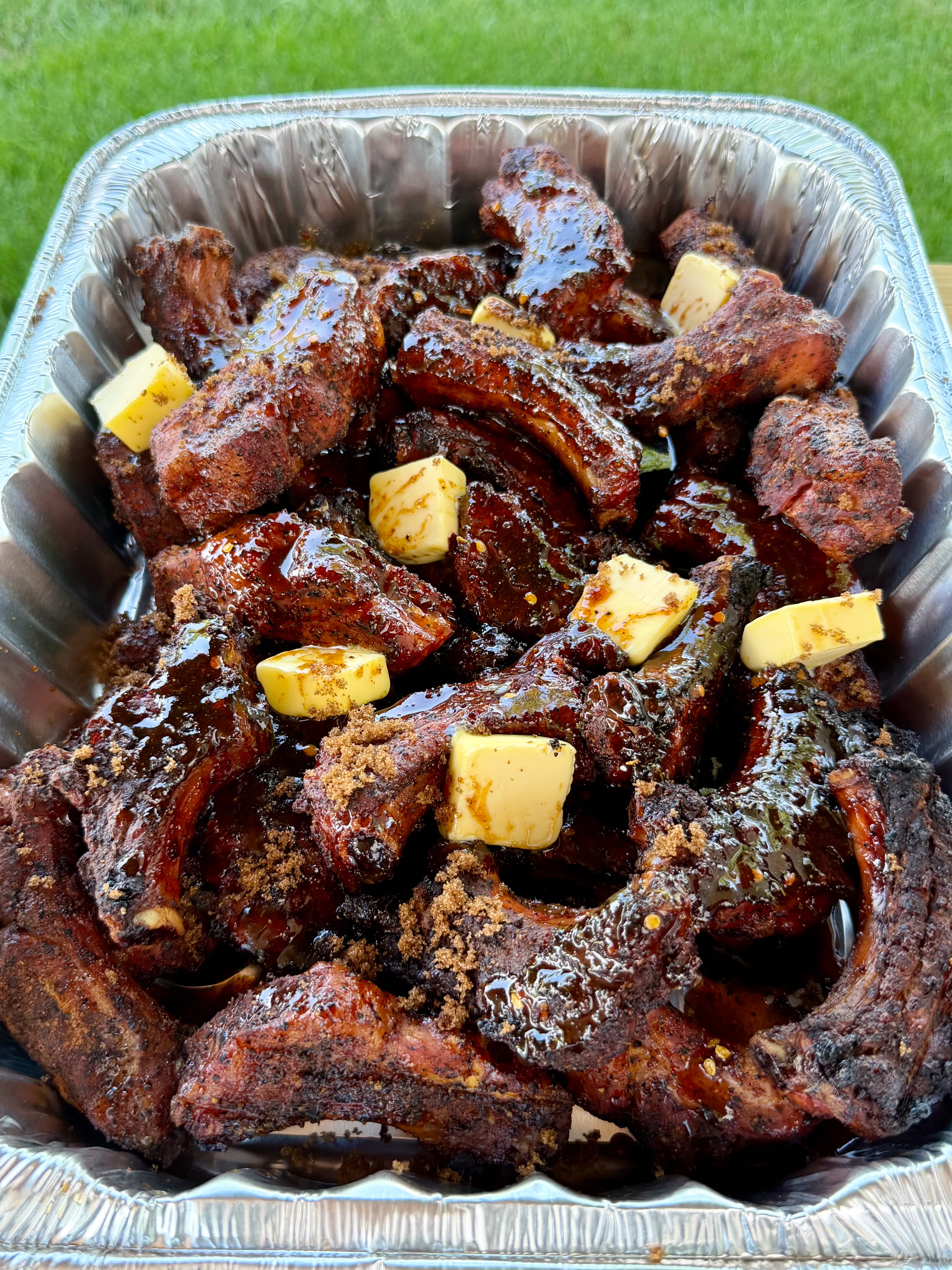 Smoked party ribs in a foil pan glazed with bbq sauce, butter pats, and brown sugar