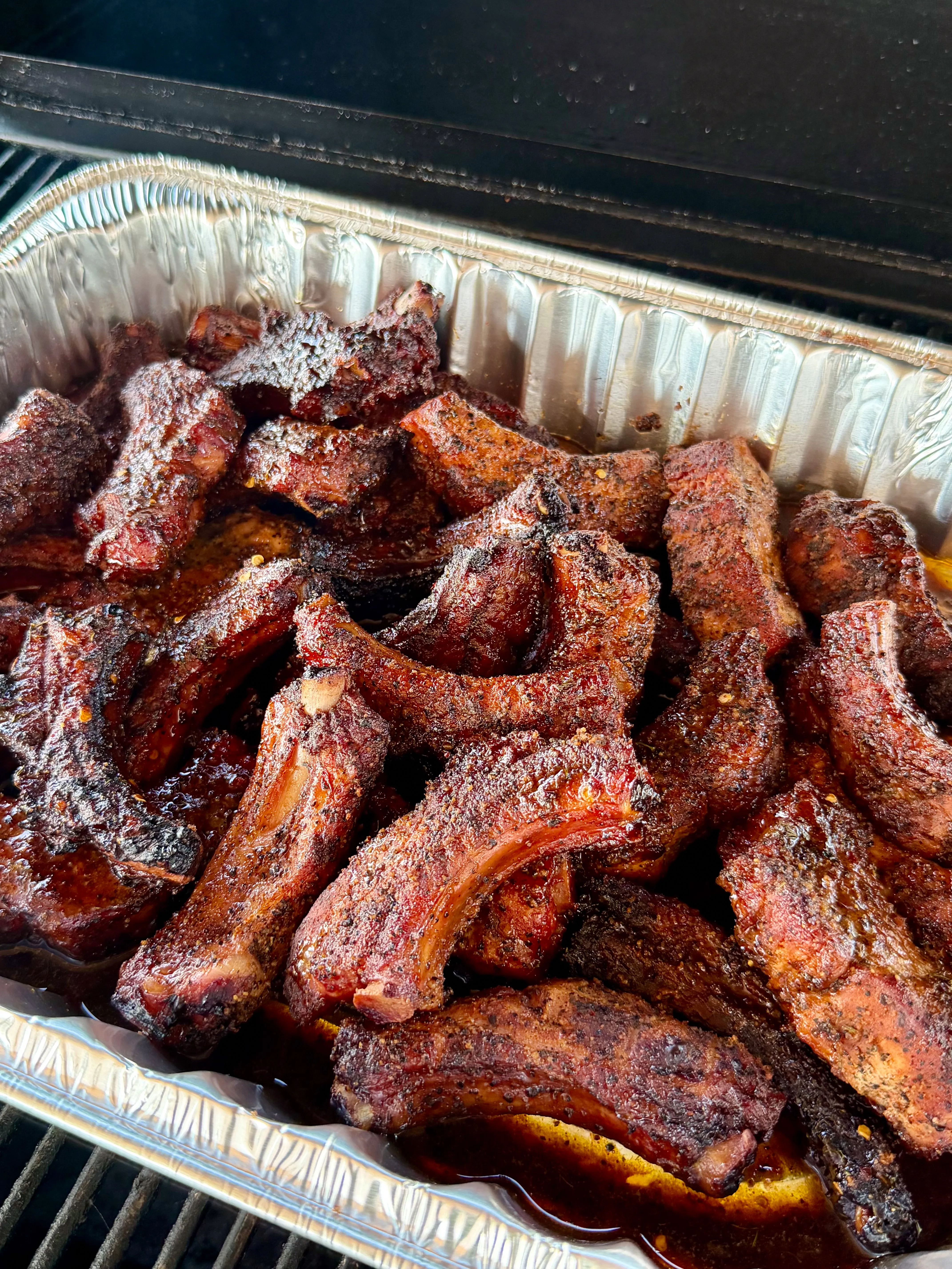 Smoked party ribs piled in an aluminum pan on a smoker grill