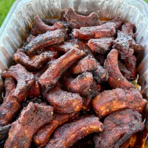 Smoked dill pickle hot honey party ribs piled high in a disposable aluminum foil pan outdoors