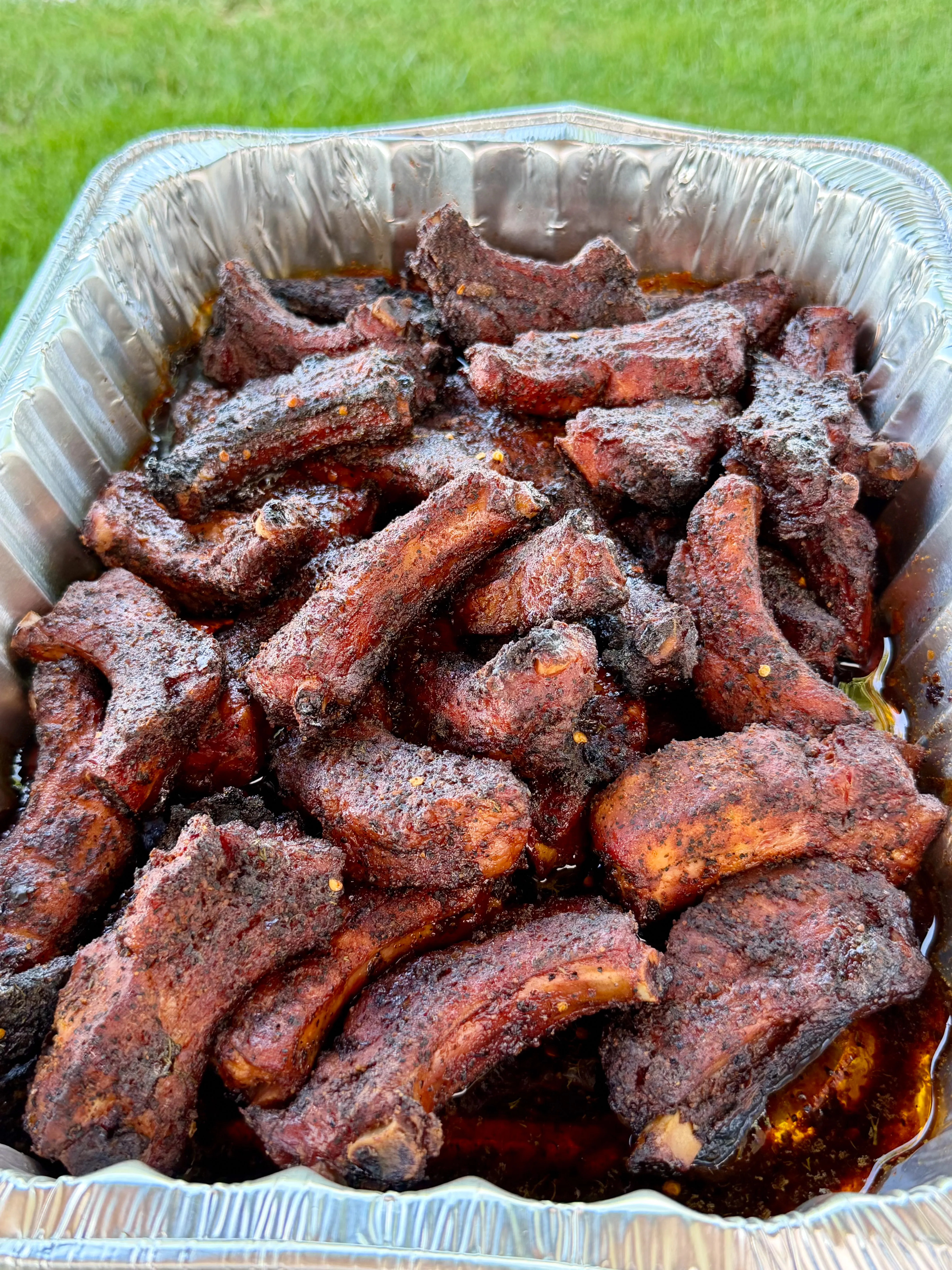 Smoked party ribs piled high in a disposable aluminum foil pan outdoors