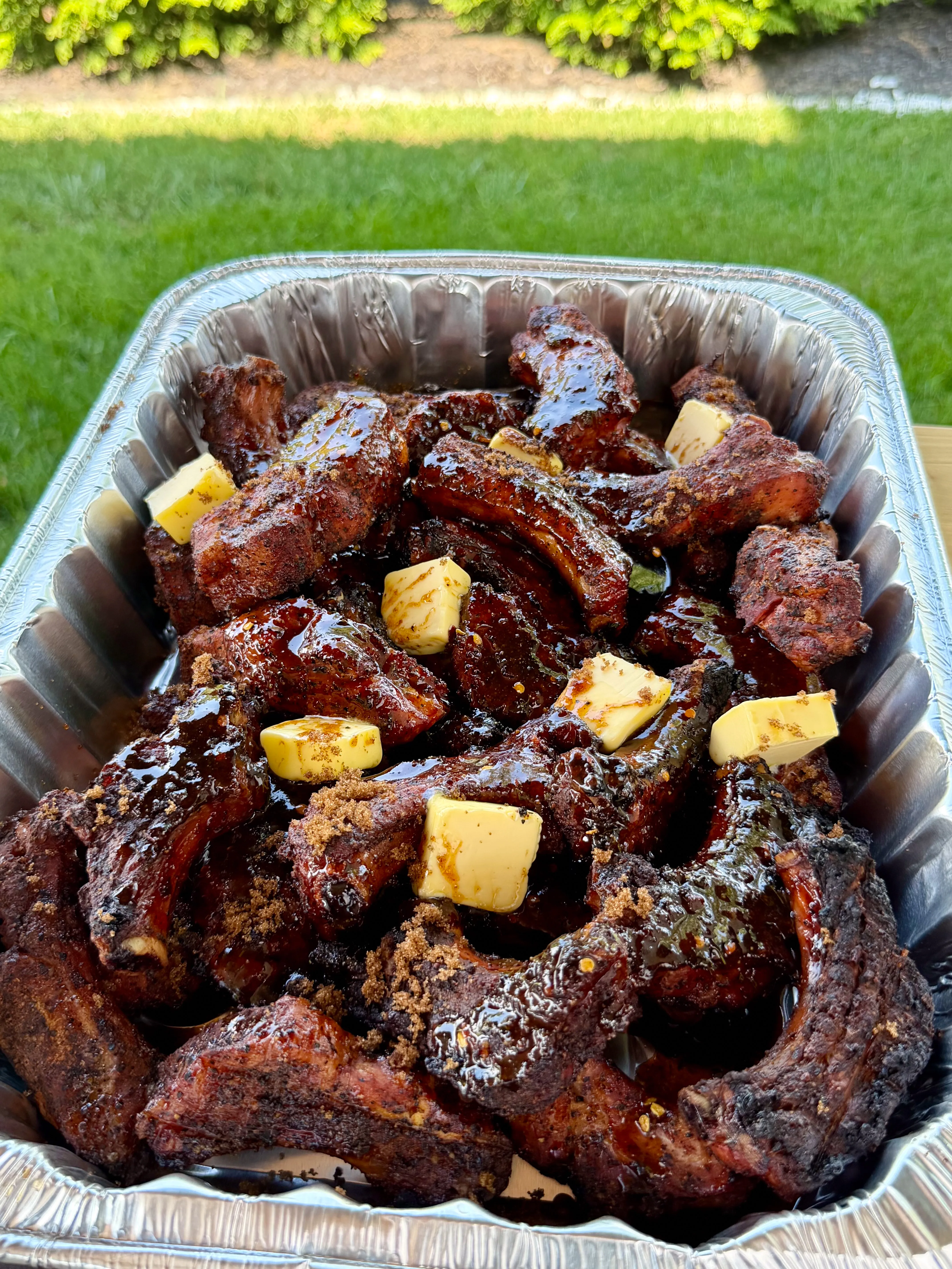 Glazed smoked party ribs with butter pats and brown sugar in an aluminum pan outdoors
