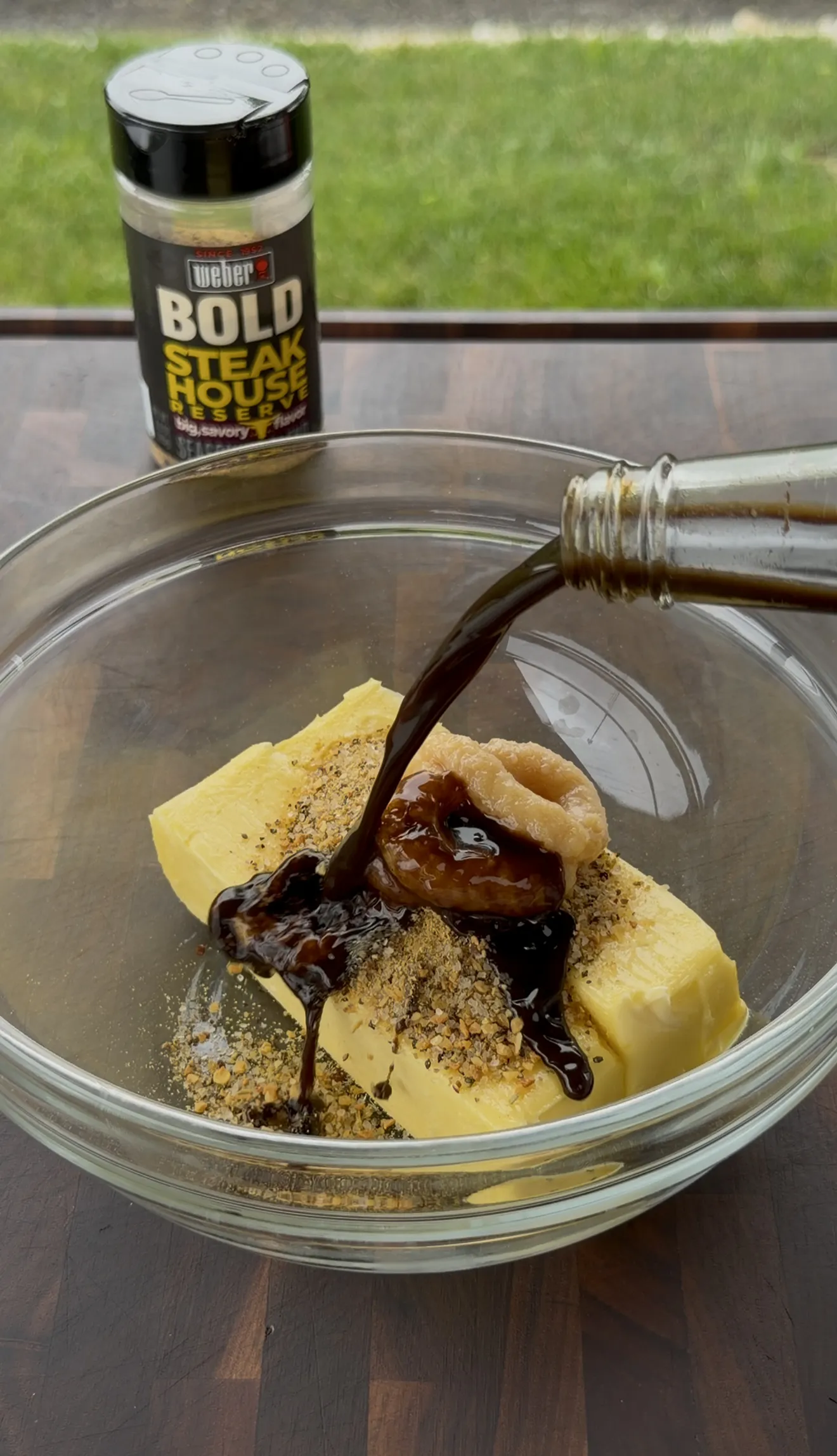 Worcestershire pouring over softened butter, roasted garlic, and seasoning in a glass bowl