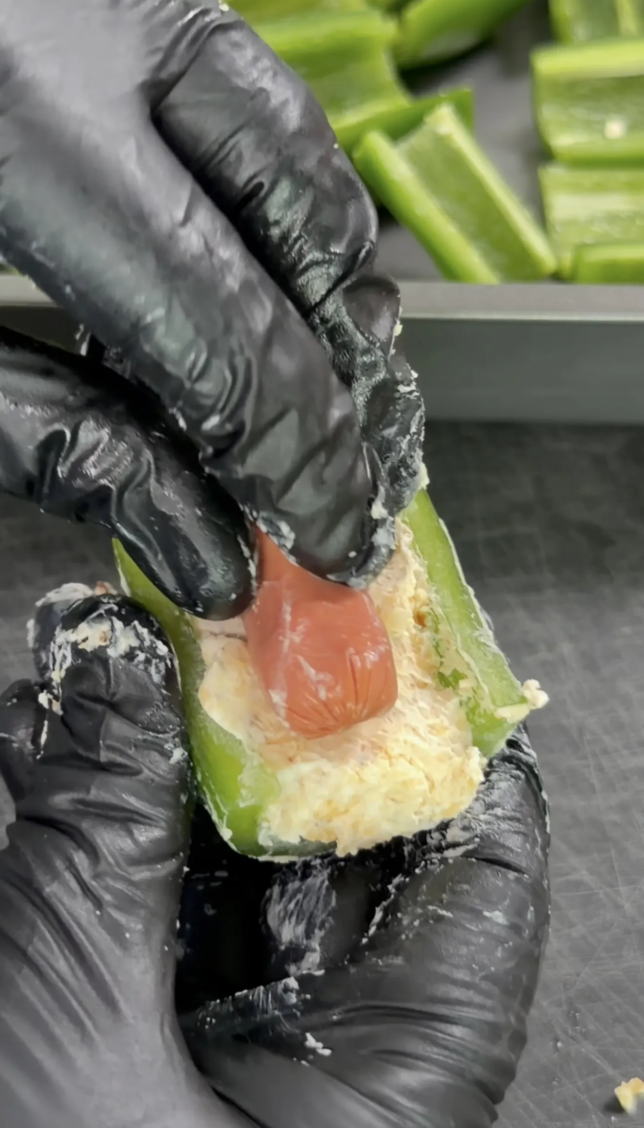 Gloved hands stuffing a halved jalapeño with cream cheese filling and a smokie for smokie bombs