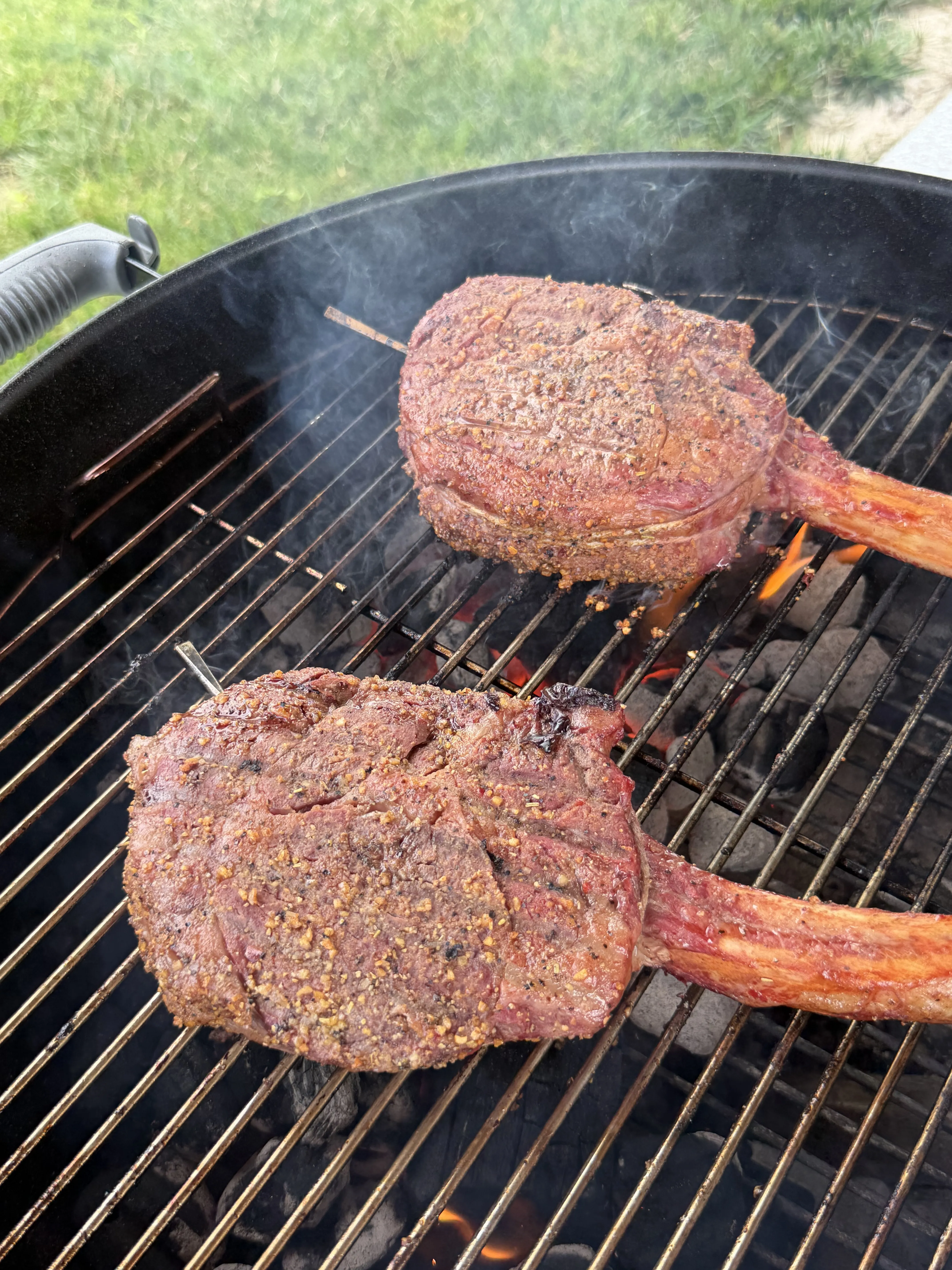 Two seasoned tomahawk ribeyes searing over hot charcoal flames