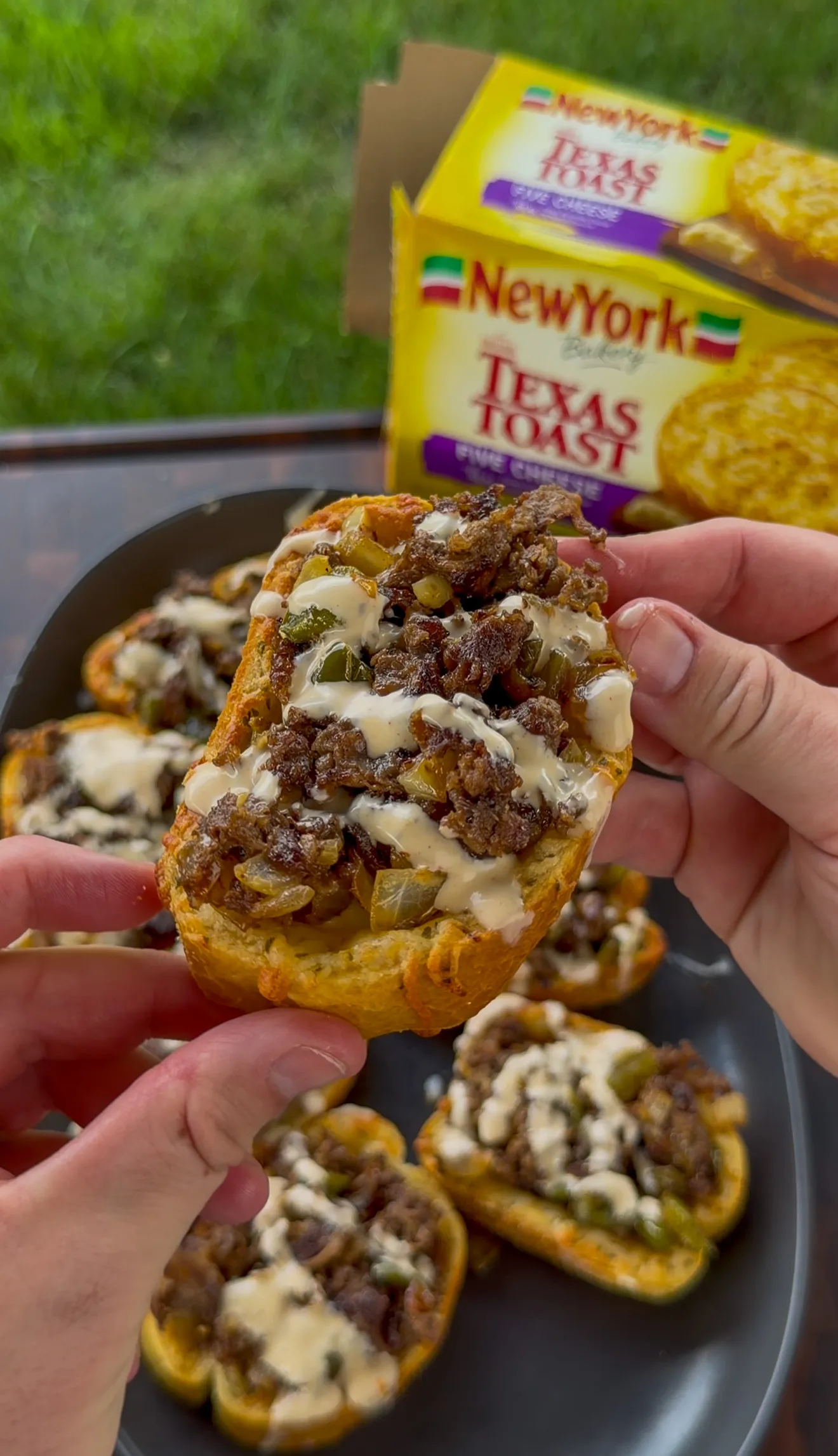 Hands holding Cheesesteak Texas Toast topped with steak, peppers, onions, and a cheese drizzle